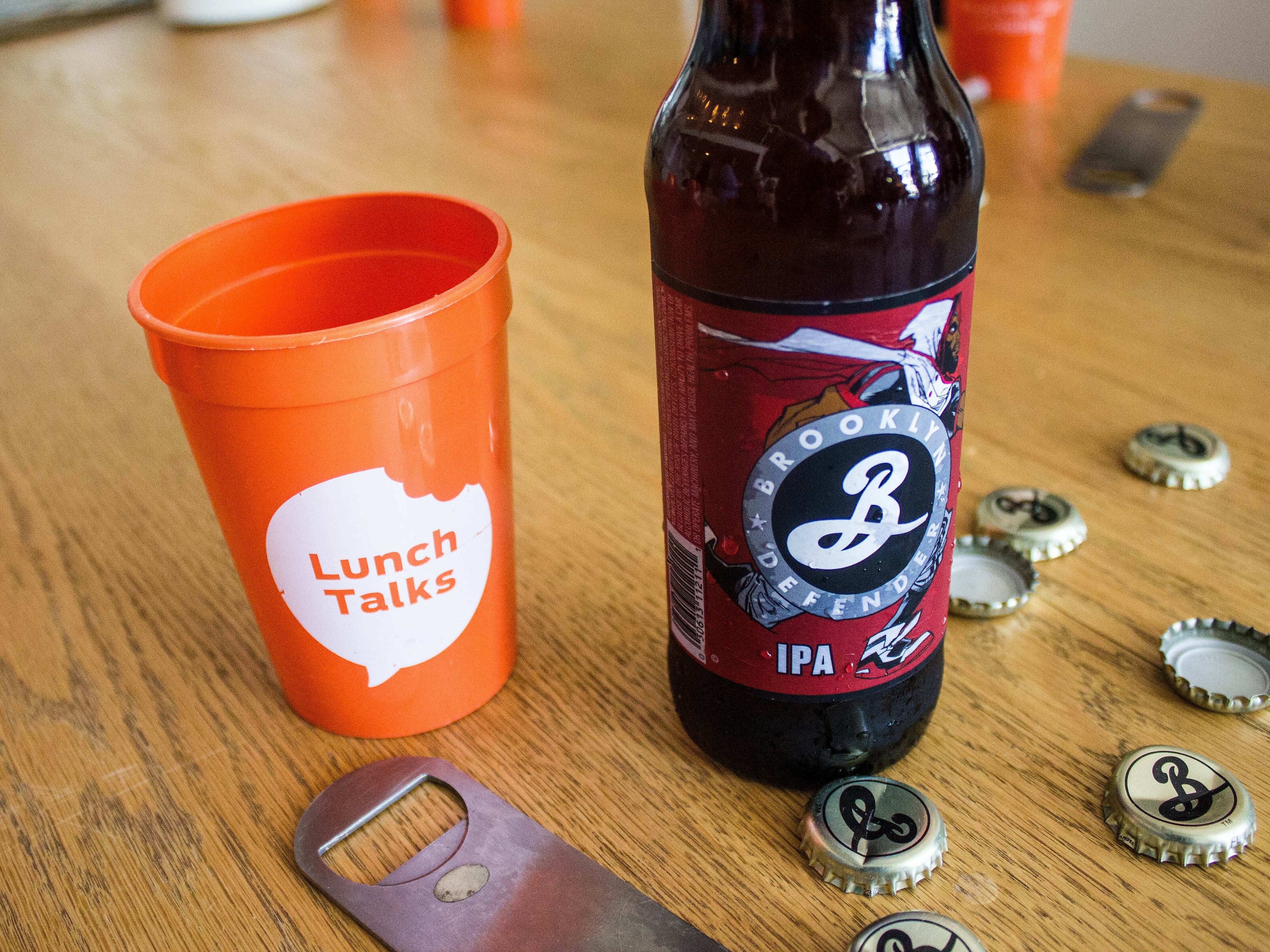 An orange cup with the words "Lunch Talks" next to a bottle of Brooklyn Defender IPA on a wooden table. Several bottle caps and an opener are scattered around the bottle.