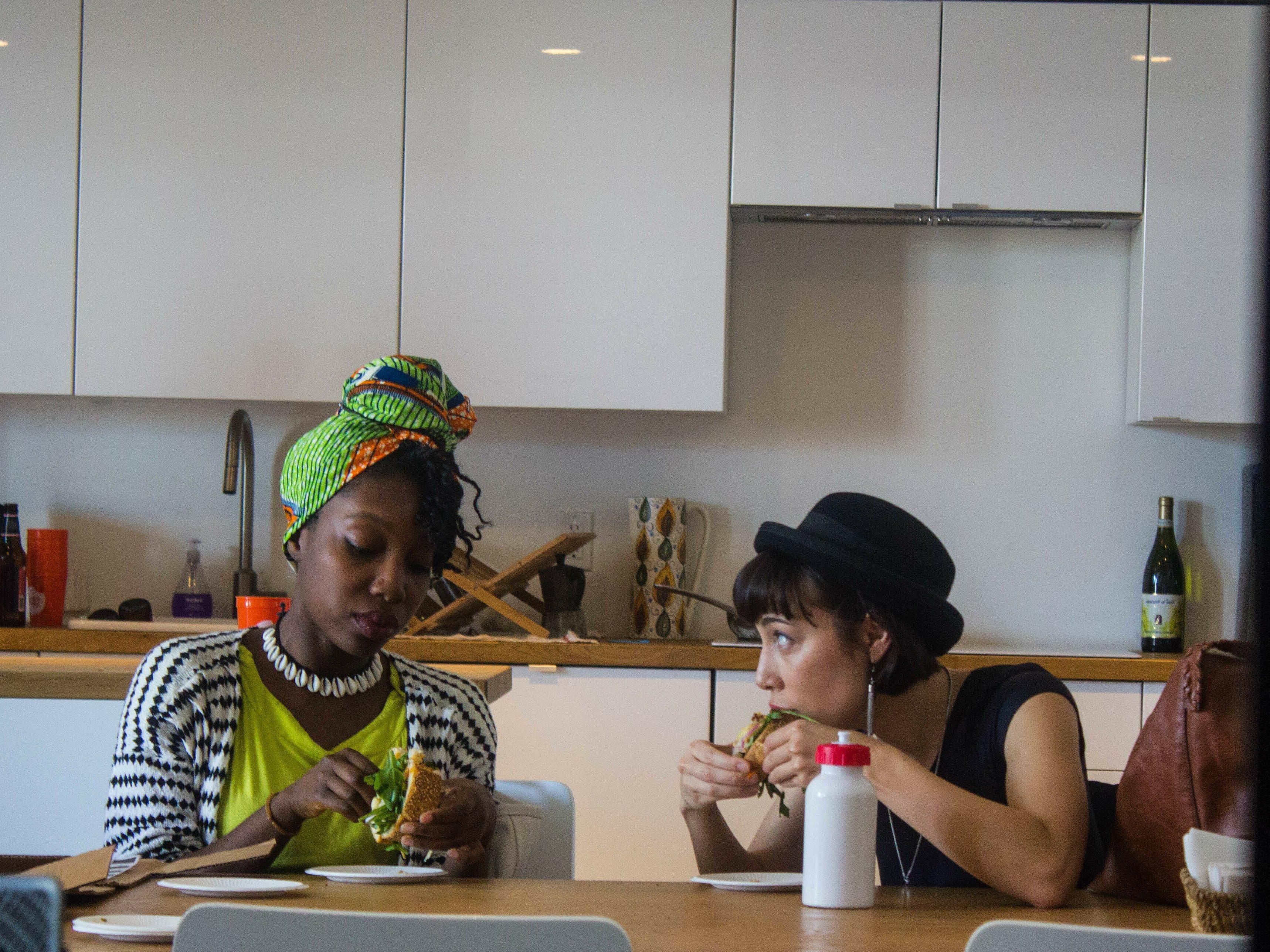 Two people are sitting at a table, eating sandwiches. One person on the left wears a green headscarf and striped cardigan, the other on the right wears a black hat. A white plastic bottle is on the table. The background features kitchen cabinets, a countertop, and utensils.