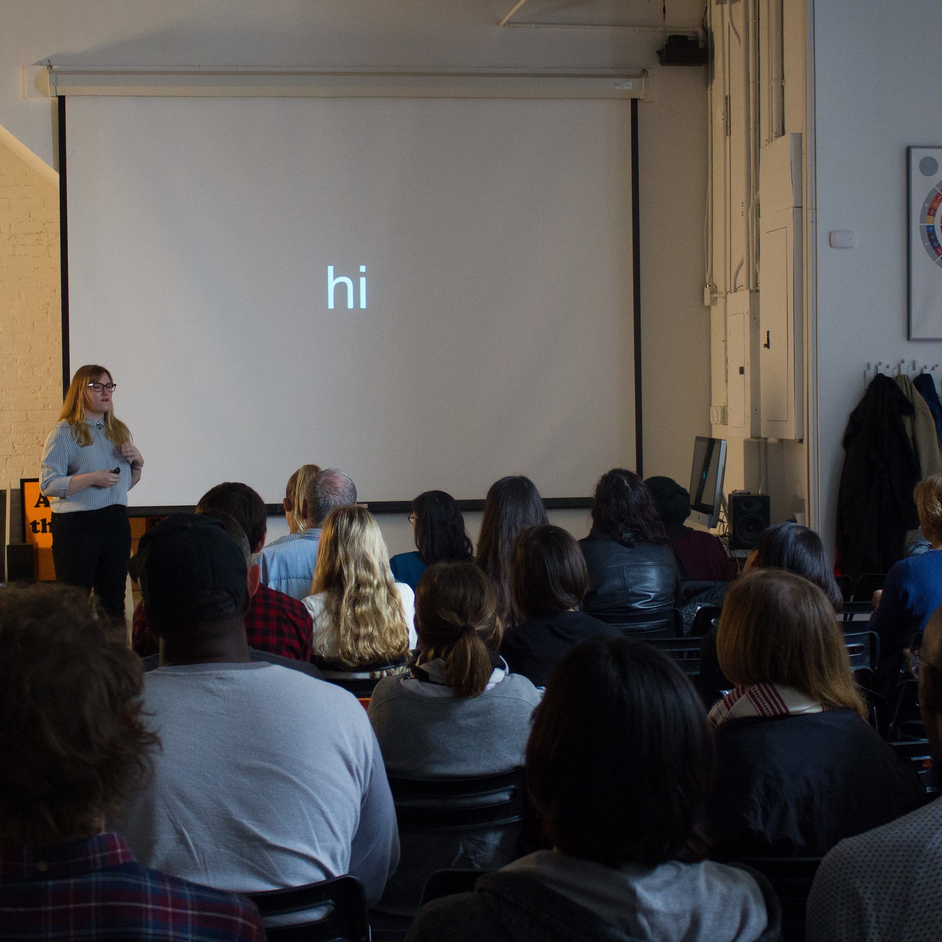 A woman stands in front of a large screen that displays the word "hi" in a room filled with seated people. She is giving a presentation in a casual, well-lit environment with various items and decorations visible around the room.
