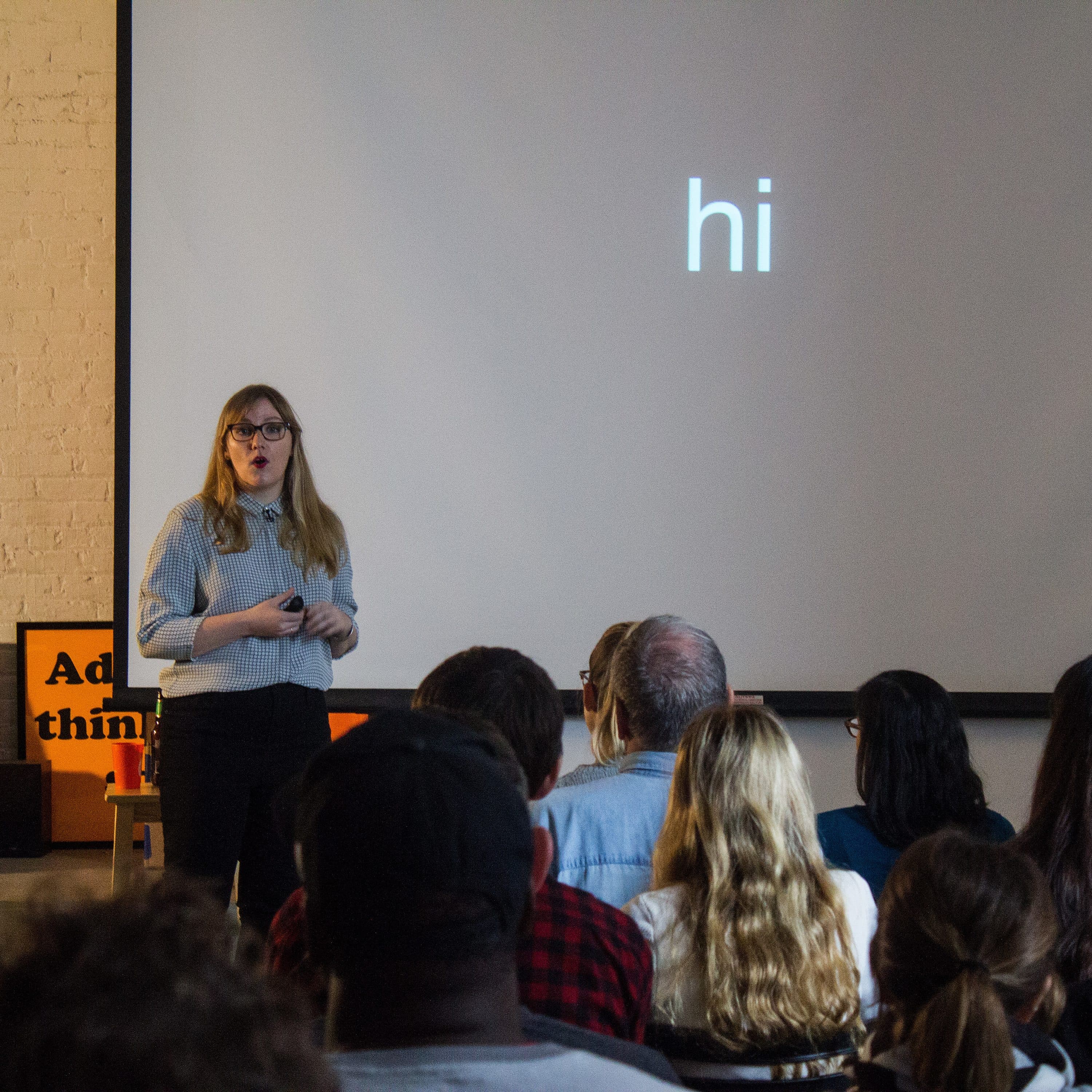 A woman stands in front of an audience, giving a presentation. The screen behind her displays the word "hi" in large, white letters. The setting appears to be an indoor venue with a relaxed, informal atmosphere. The audience is seated and attentively listening.