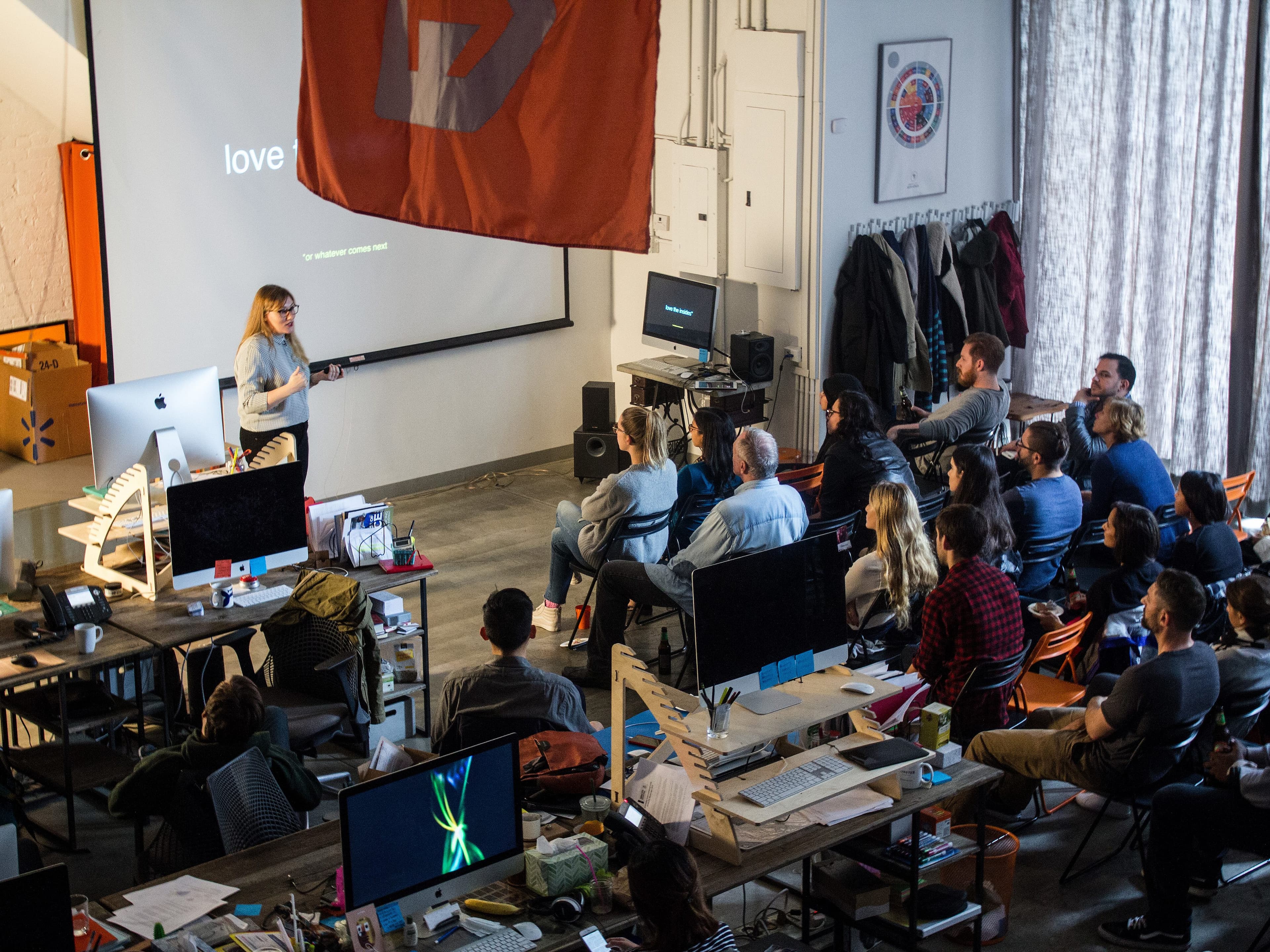 A woman stands in front of a projector screen giving a presentation to a seated audience in a cluttered office space. The screen displays the word "love" and a red flag hangs from the ceiling. People are attentively watching and taking notes.
