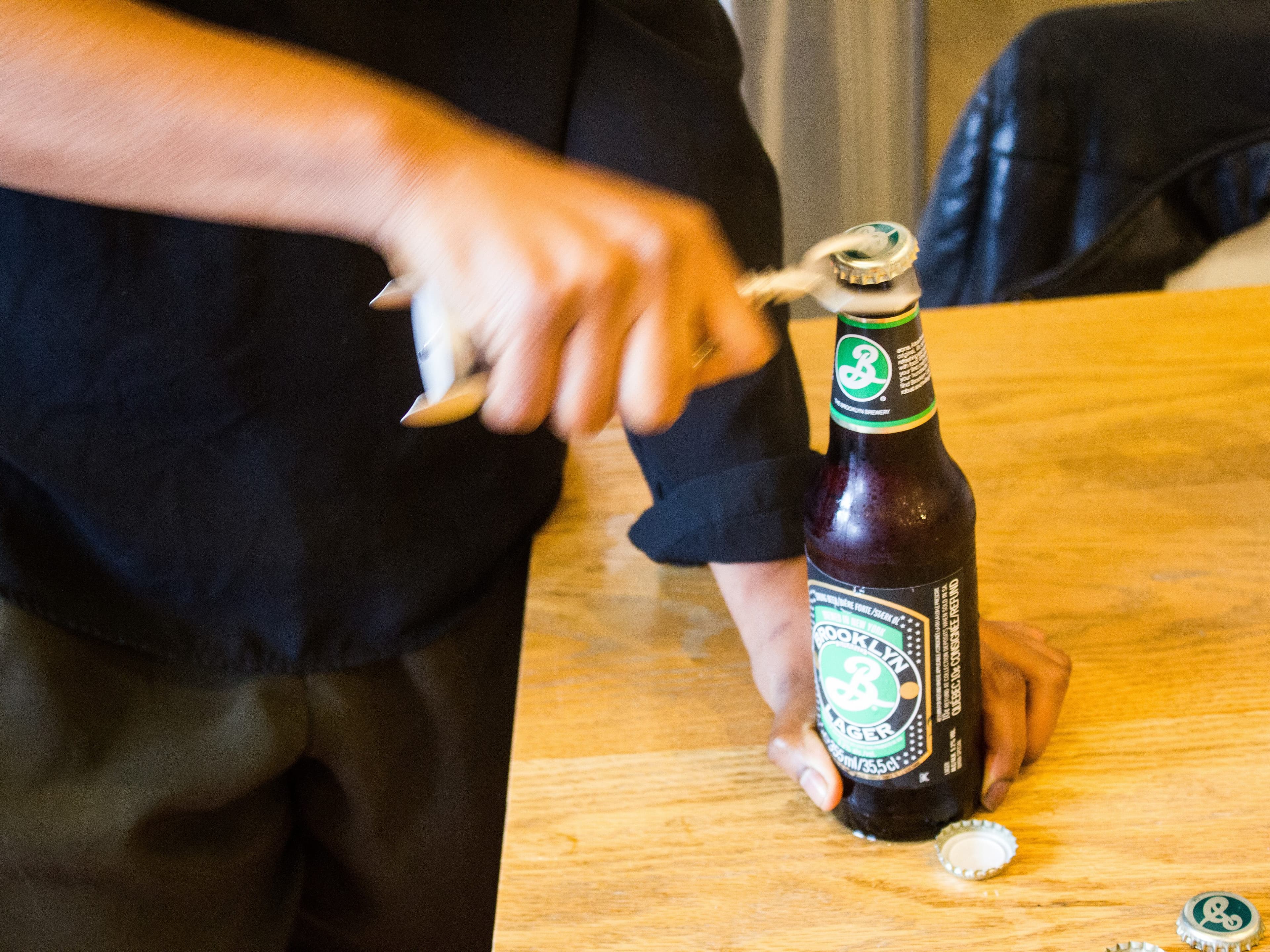 A person is opening a Brooklyn Brewery beer bottle with a bottle opener. The person holds the bottle with one hand on a wooden table, while using the other hand to open it. Several bottle caps are scattered on the table.