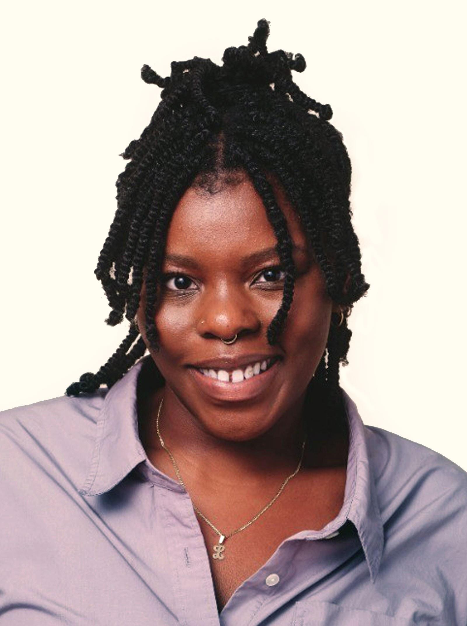 A person with braided hair smiles at the camera. They are wearing a purple button-up shirt and a necklace with a small pendant. The background is a plain light color.