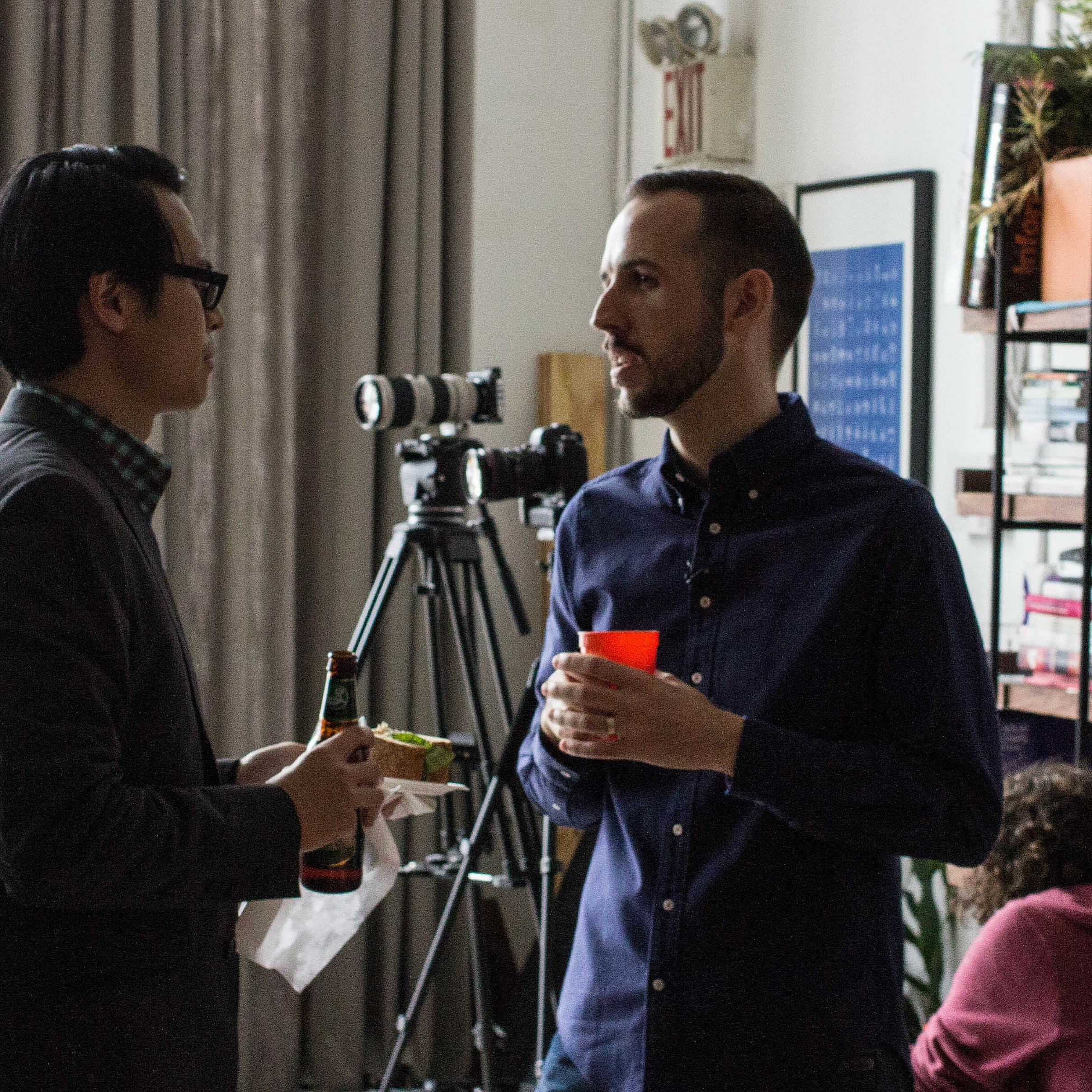 Two people in conversation indoors. One, holding a drink and snacks, wears glasses and a casual blazer. The other, holding a red cup, has a beard and is dressed in a dark shirt. A camera on a tripod and bookshelves in the background suggest a creative or informal setting.
