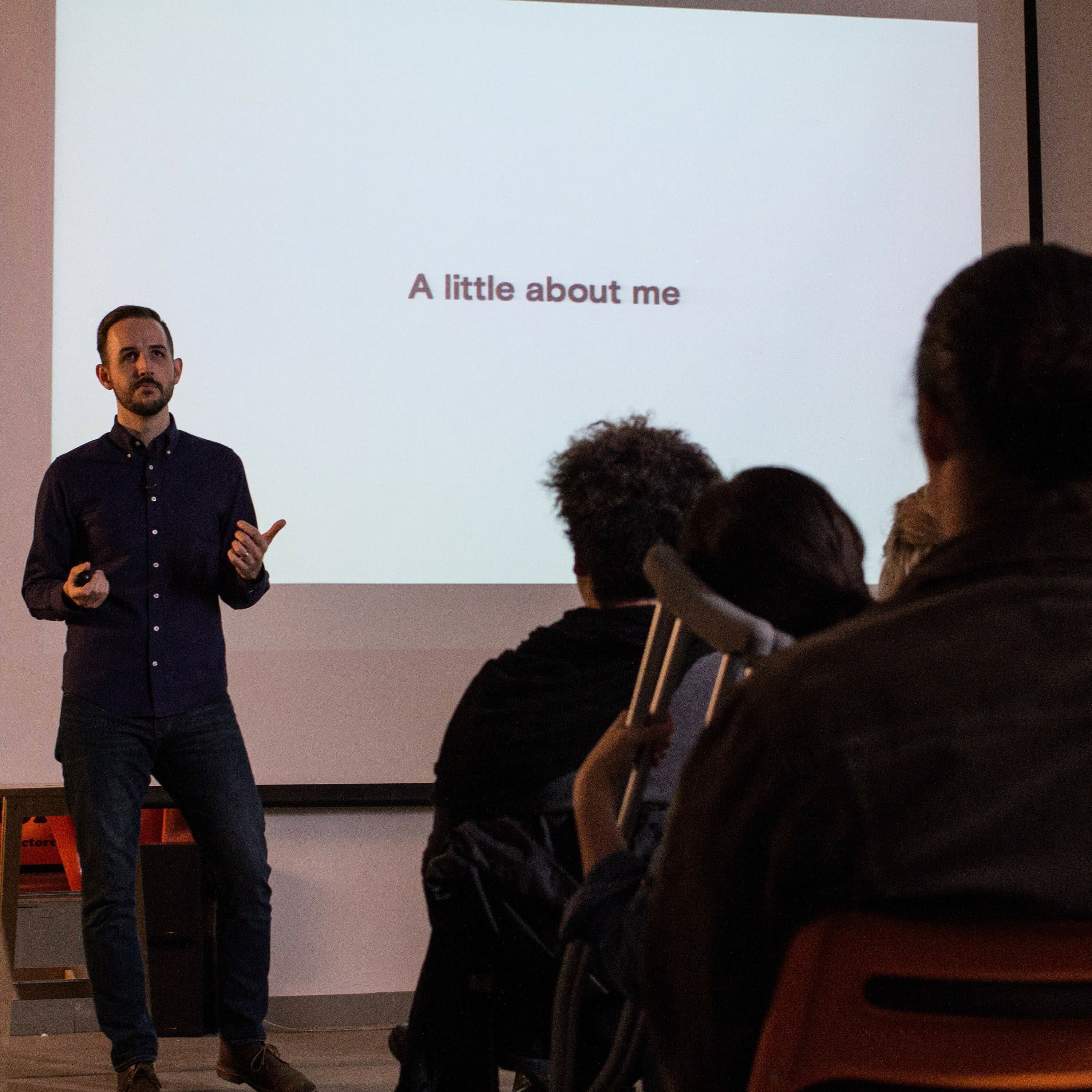 A person stands in front of a screen displaying the text "A little about me," giving a presentation to an audience seated in a room. The presenter, dressed in a dark shirt and jeans, gestures with one hand while holding a remote in the other.