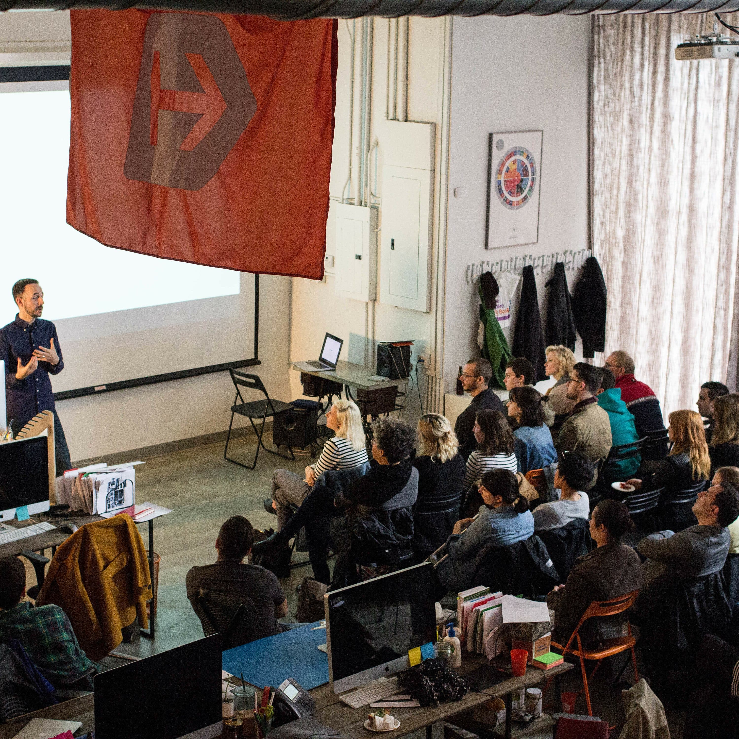 A speaker is giving a presentation to a seated audience in a modern office space. A large red flag with a logo hangs overhead, and several computer monitors and office equipment are visible in the foreground. The audience is engaged and attentive.