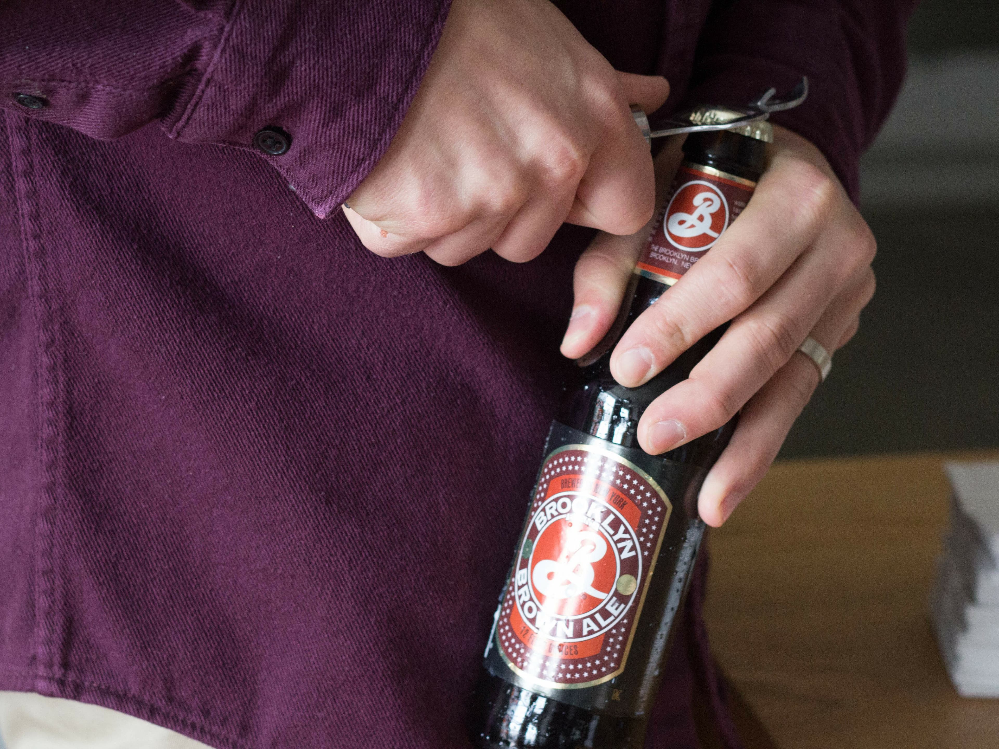 A person wearing a maroon shirt is opening a Brooklyn Brewery brand beer bottle with a metal bottle opener. The person is holding the bottle cap and the opener with one hand and the bottle with the other. The bottle label is clearly visible.