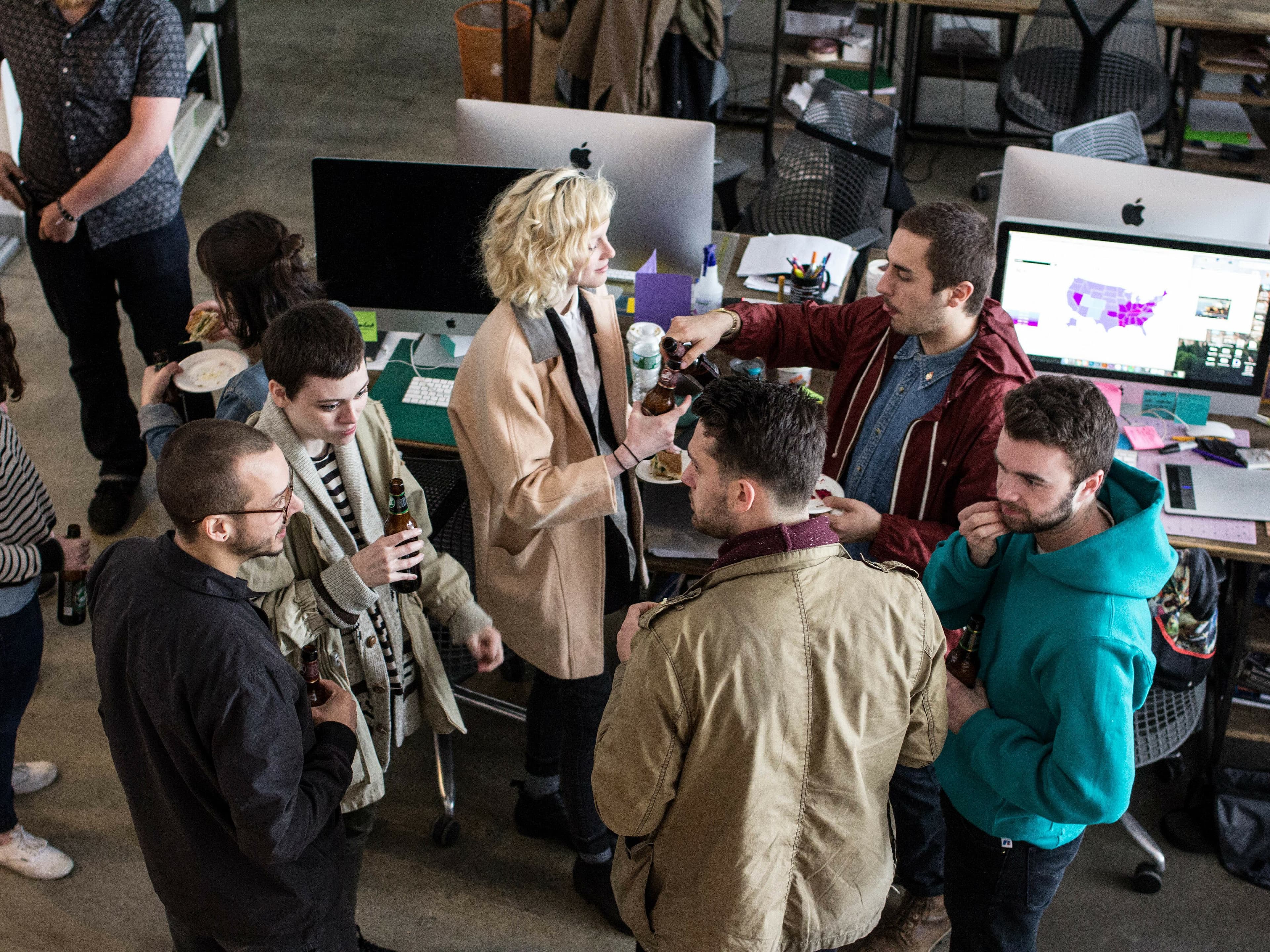 A group of people gather in an office environment, drinking and socializing near a couple of desks with iMac computers. Some are holding drinks, and one person appears to be pouring a drink for another. The atmosphere looks casual and friendly.
