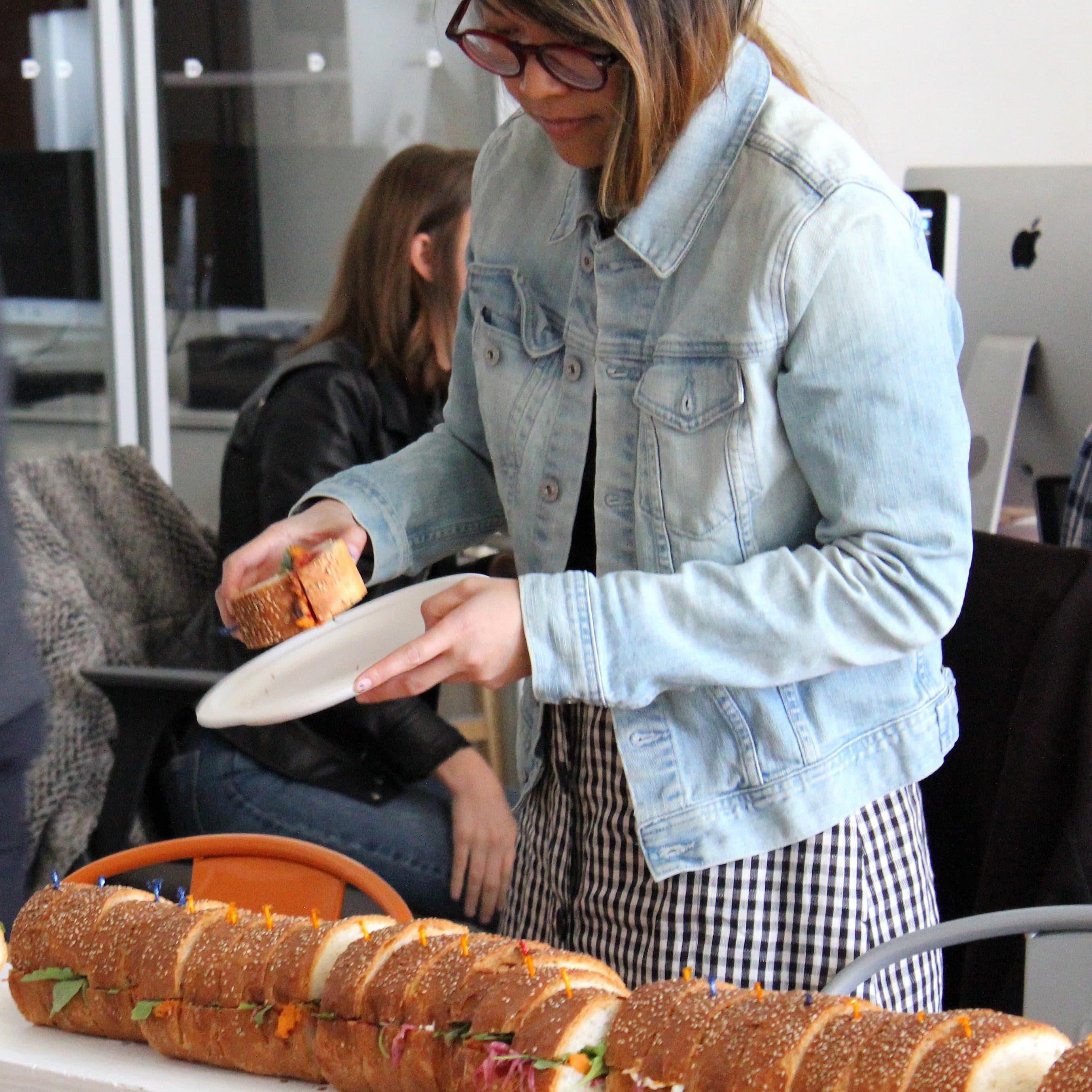 A person wearing glasses and a denim jacket is serving themselves a sandwich from a large sub that extends off the table. They hold a plate in one hand and use the other to pick up a sandwich section. In the background, other people are seated at desks with computers.