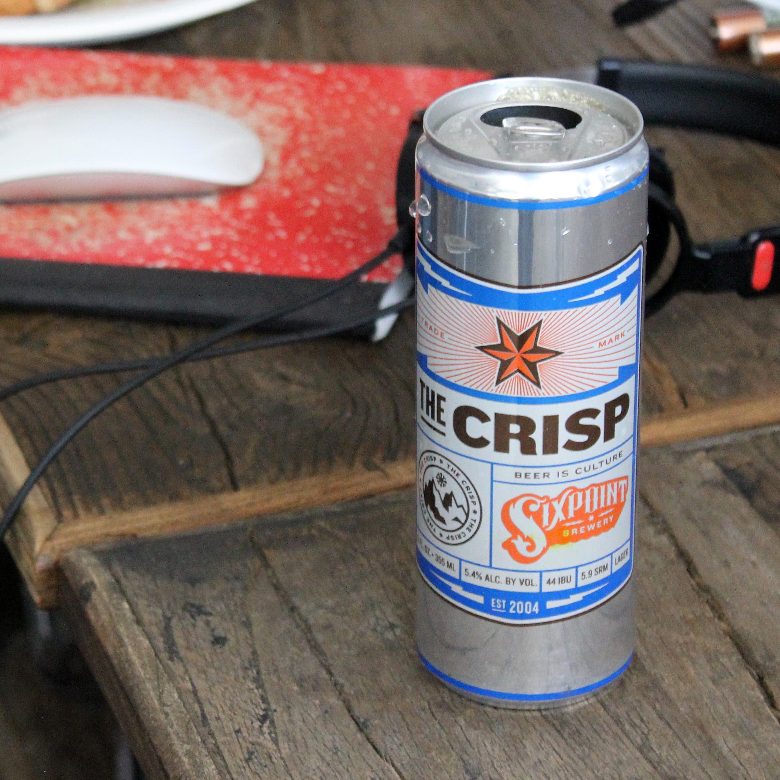 A can of Sixpoint "The Crisp" beer rests on a wooden table near a red notebook, white computer mouse, and a pair of black earbuds. The can features a blue and silver design with a star logo and the phrase "Beer is Culture.