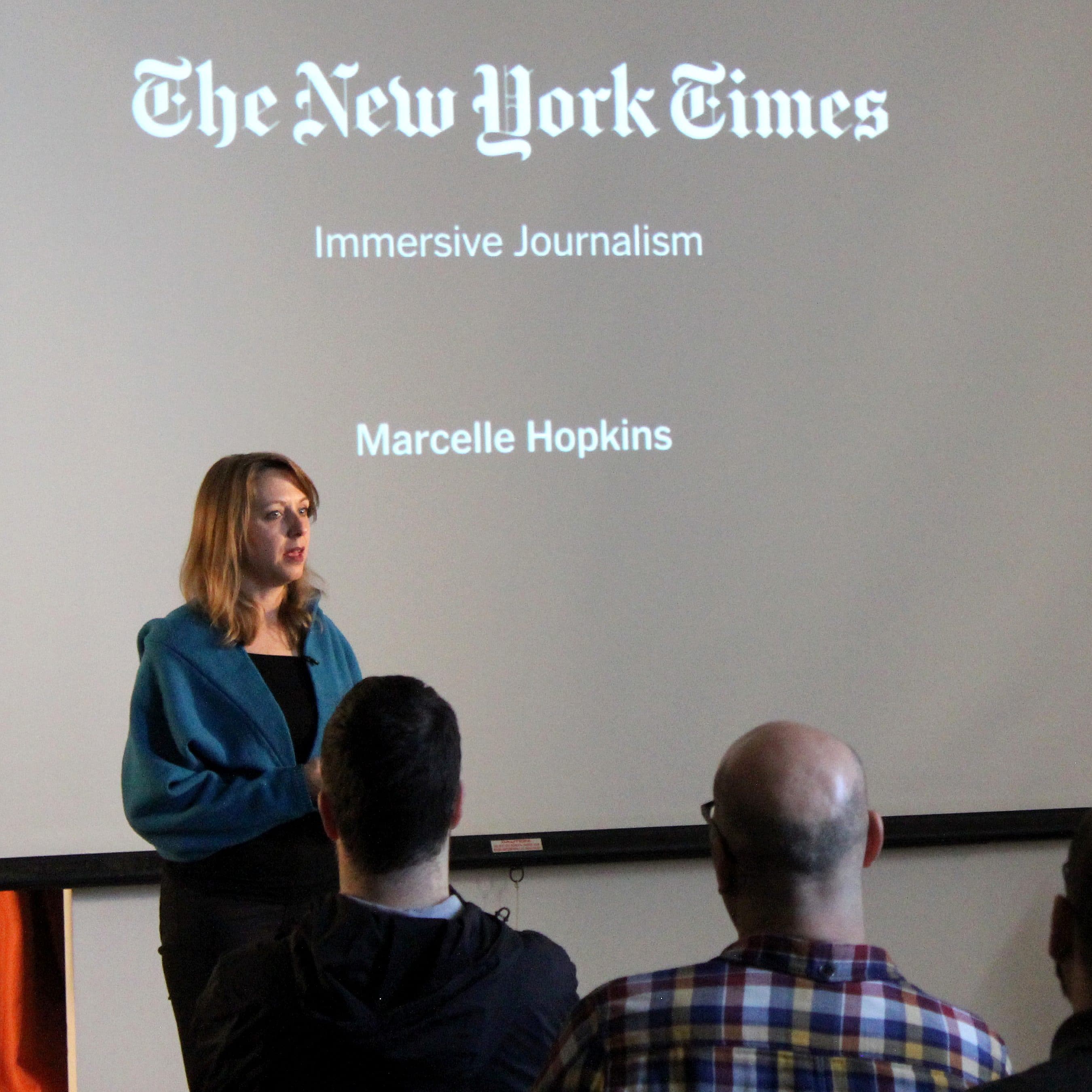 A woman stands and speaks in front of a projected presentation slide titled "The New York Times Immersive Journalism" with the name "Marcelle Hopkins" below it. She is addressing an audience of seated people.