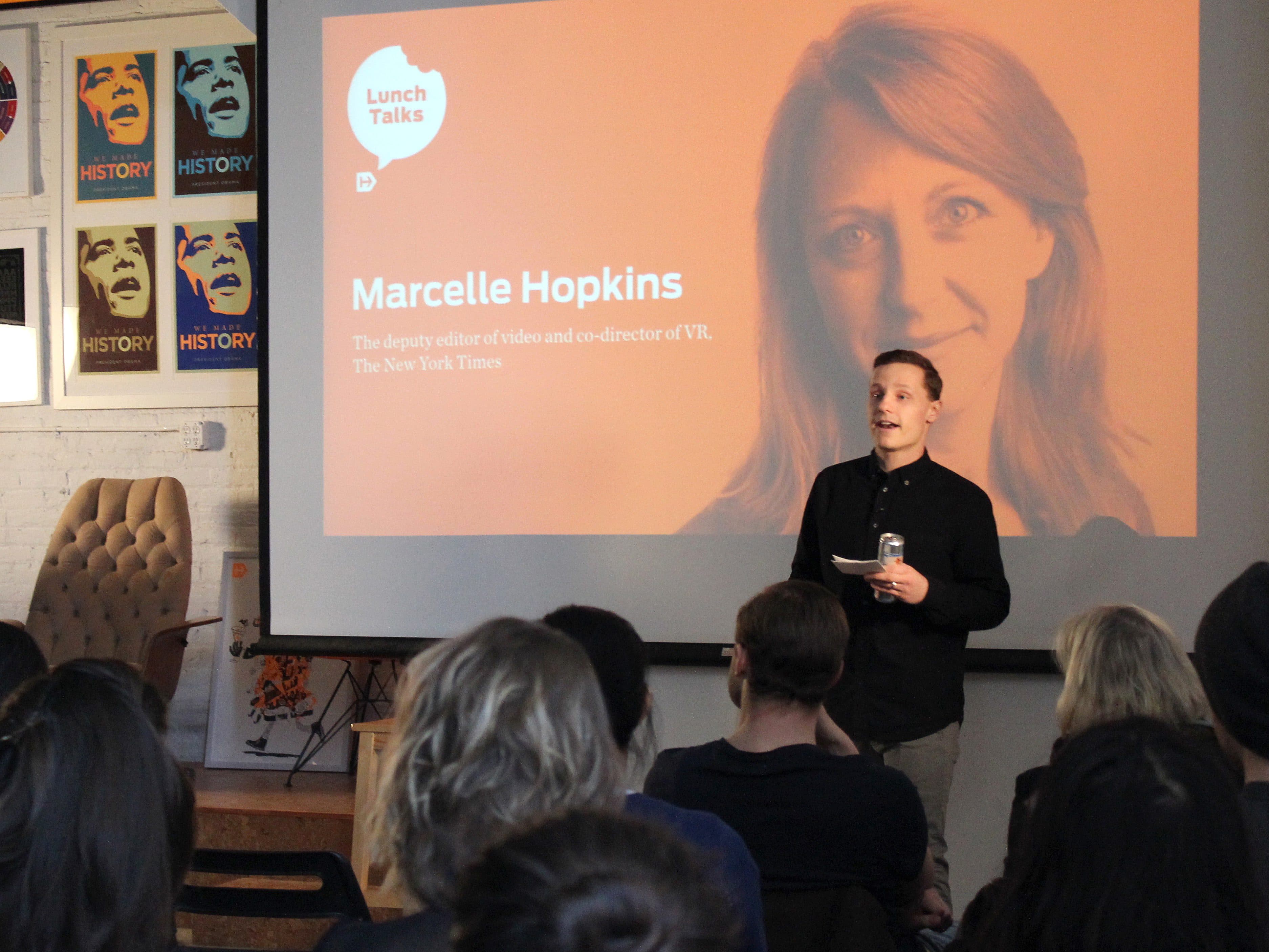 A person stands in front of an audience, holding a glass and speaking, with a presentation slide projected behind them. The slide features a person's photo with the name "Marcelle Hopkins," and their title as "The deputy editor of video and co-director of VR, The New York Times.