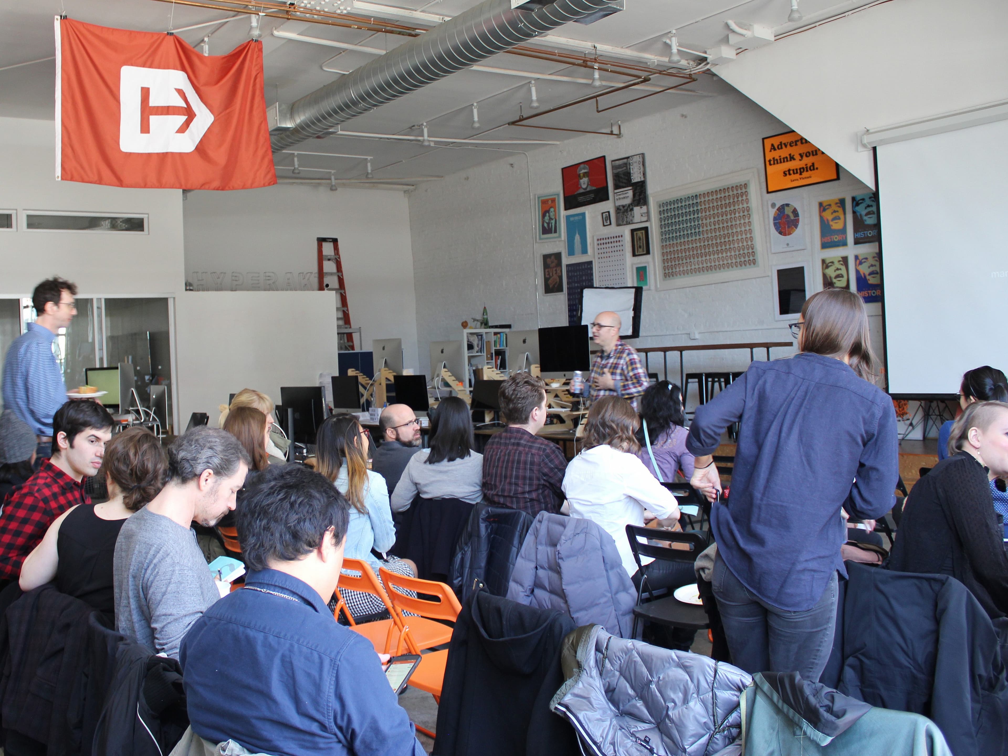 A group of people sits in a classroom with a large orange flag bearing a white "H" hanging from the ceiling. The room has a projector screen, posters on the wall, and a few people standing or walking around. Some attendees are talking, and most are facing toward the front.