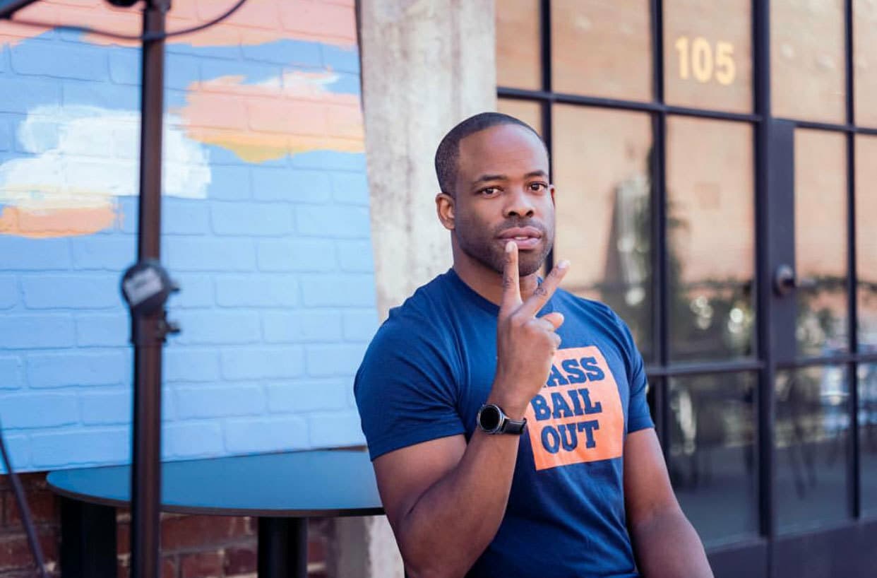 A man wearing a navy blue t-shirt with orange lettering that reads "PASS BAIL OUT" is seated outside. He is holding up a peace sign with his right hand. Behind him is a painted brick wall and a black door with the number 105.