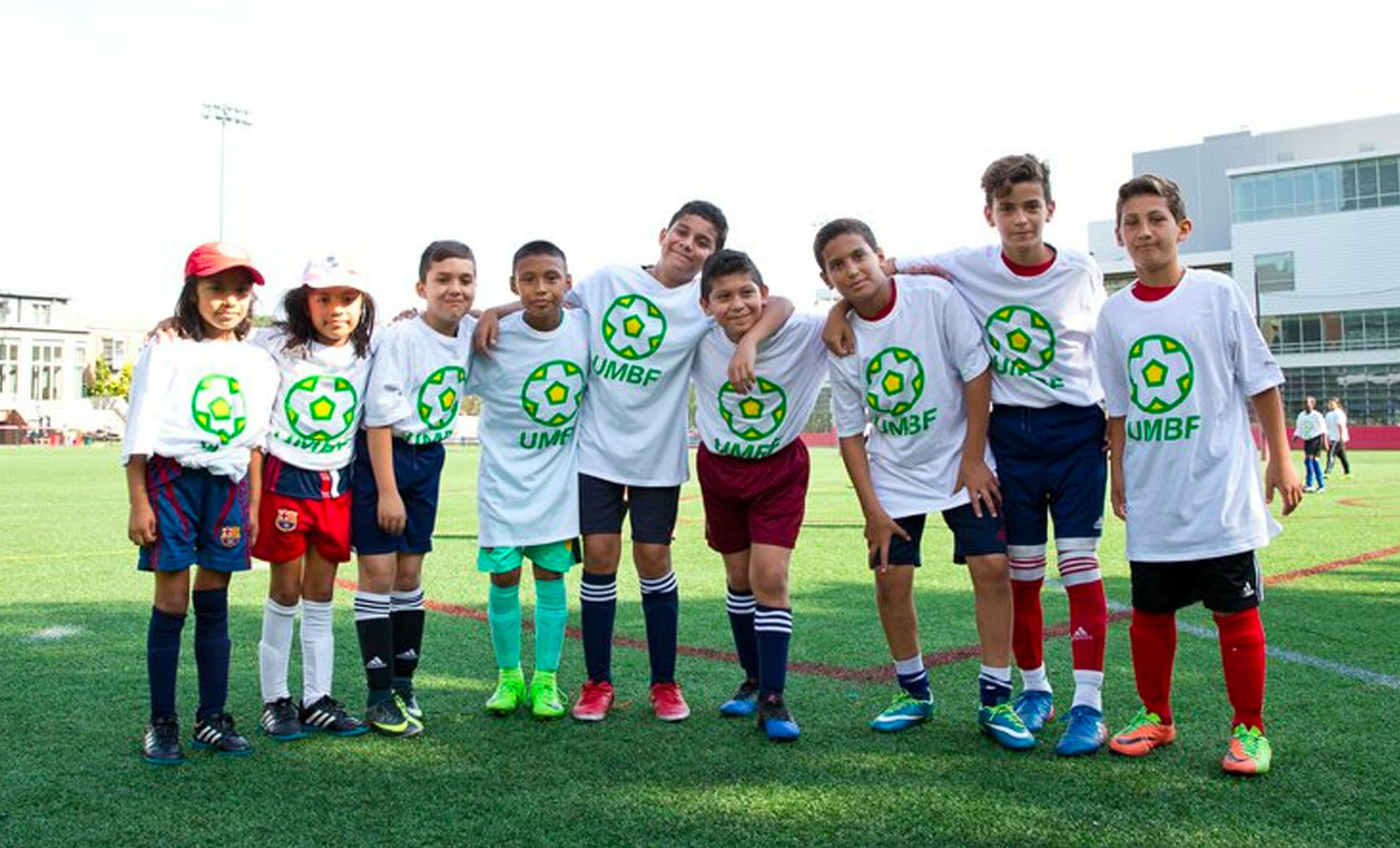 A group of nine kids standing in a row on a soccer field, each wearing a white T-shirt with a green soccer ball logo and the letters "UMBF" printed on the front. They smile and pose with arms around each other on a sunny day.