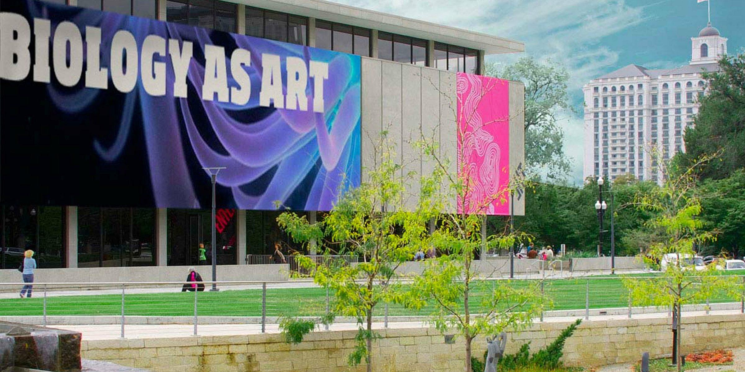 The image shows a modern building with large banners on its facade, one reading "BIOLOGY AS ART." The area in front has a landscaped garden with trimmed grass, trees, and a fountain. In the background, a tall historic building is visible against a blue sky.