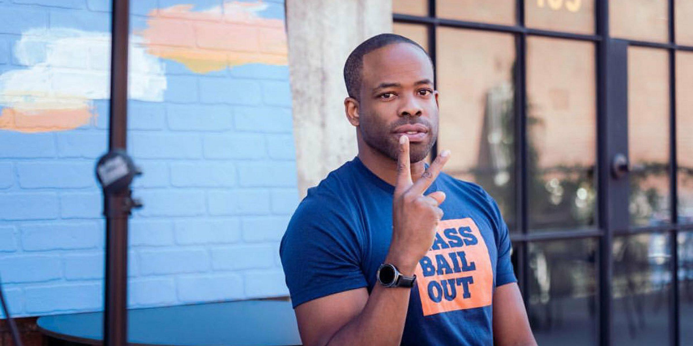 A man wearing a navy blue t-shirt with orange lettering that reads "PASS BAIL OUT" is seated outside. He is holding up a peace sign with his right hand. Behind him is a painted brick wall and a black door with the number 105.