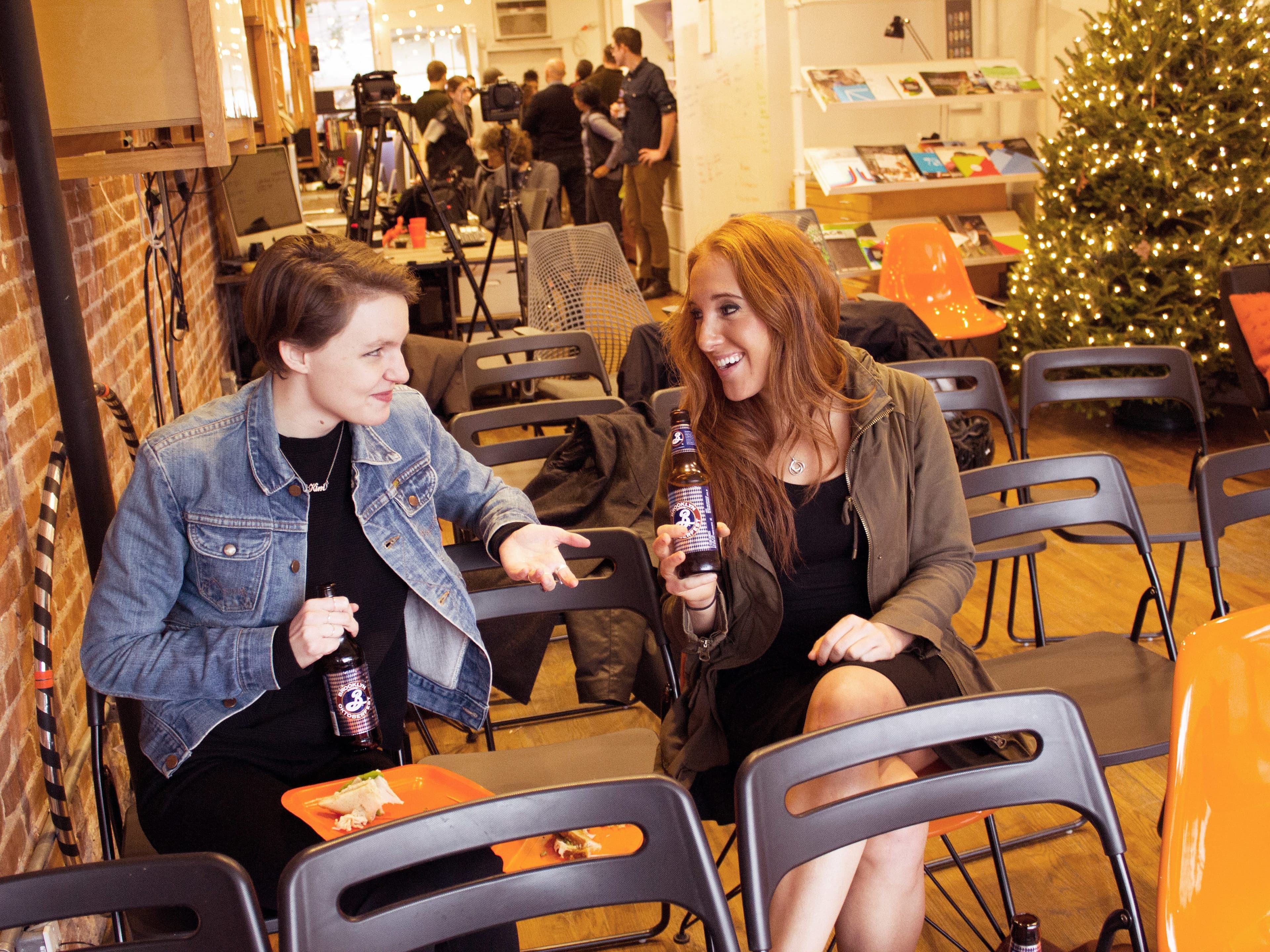 Two women are sitting on black folding chairs in a room with holiday decorations and string lights. They are smiling and holding bottles, engaging in a lively conversation. One is wearing a denim jacket, and the other is in a green jacket. A Christmas tree is in the background.