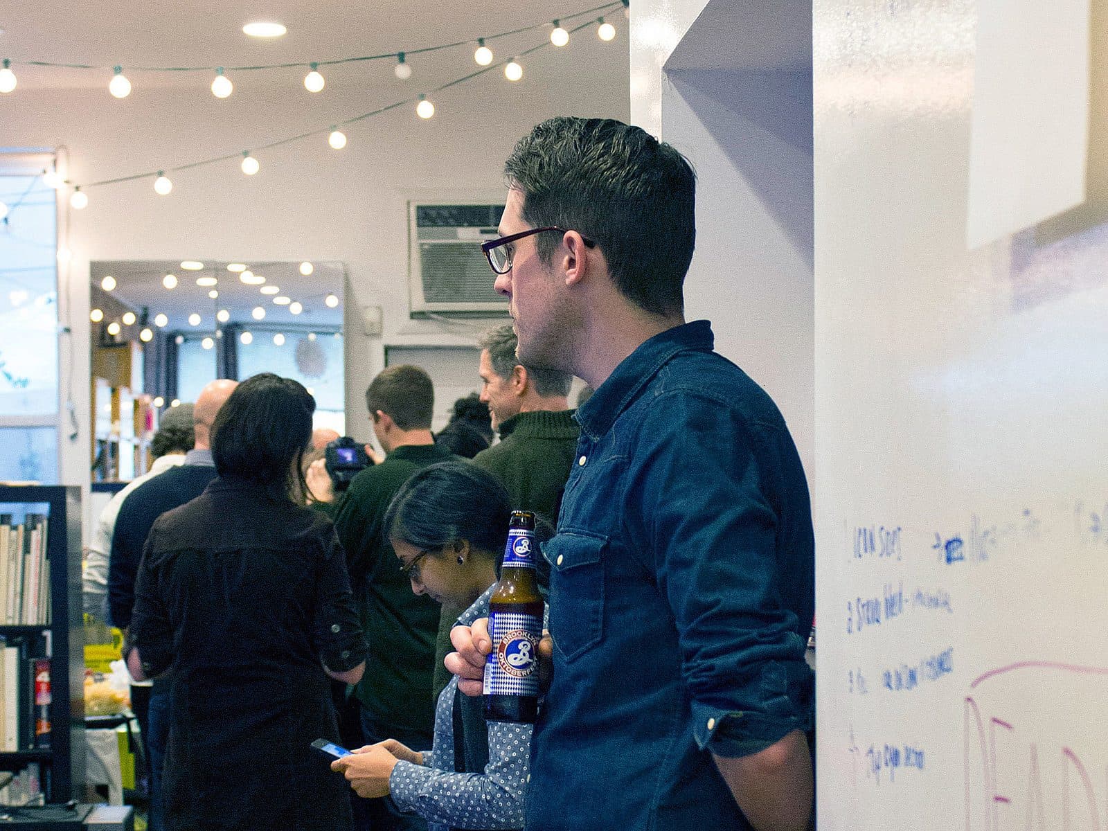 A man in glasses is holding a beer bottle, leaning against a wall in a well-lit room with string lights. Several people are gathered around, some chatting and some looking at their phones. Bookshelves and a window can be seen in the background.