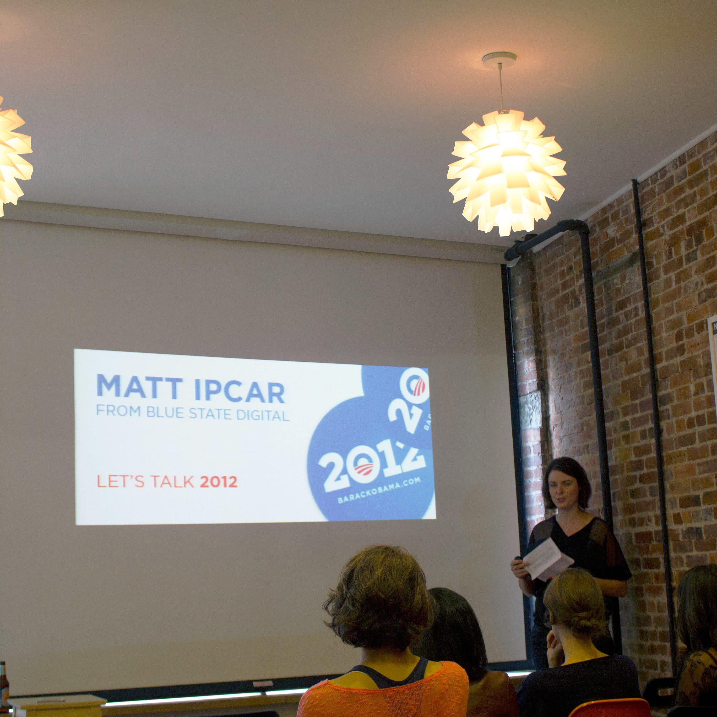 A man and a woman are presenting in front of an audience. The woman is holding a paper, while the man stands beside a projection screen that displays the text, "Matt Ipcar from Blue State Digital, Let's Talk 2012." The room has brick walls and hanging white lights.