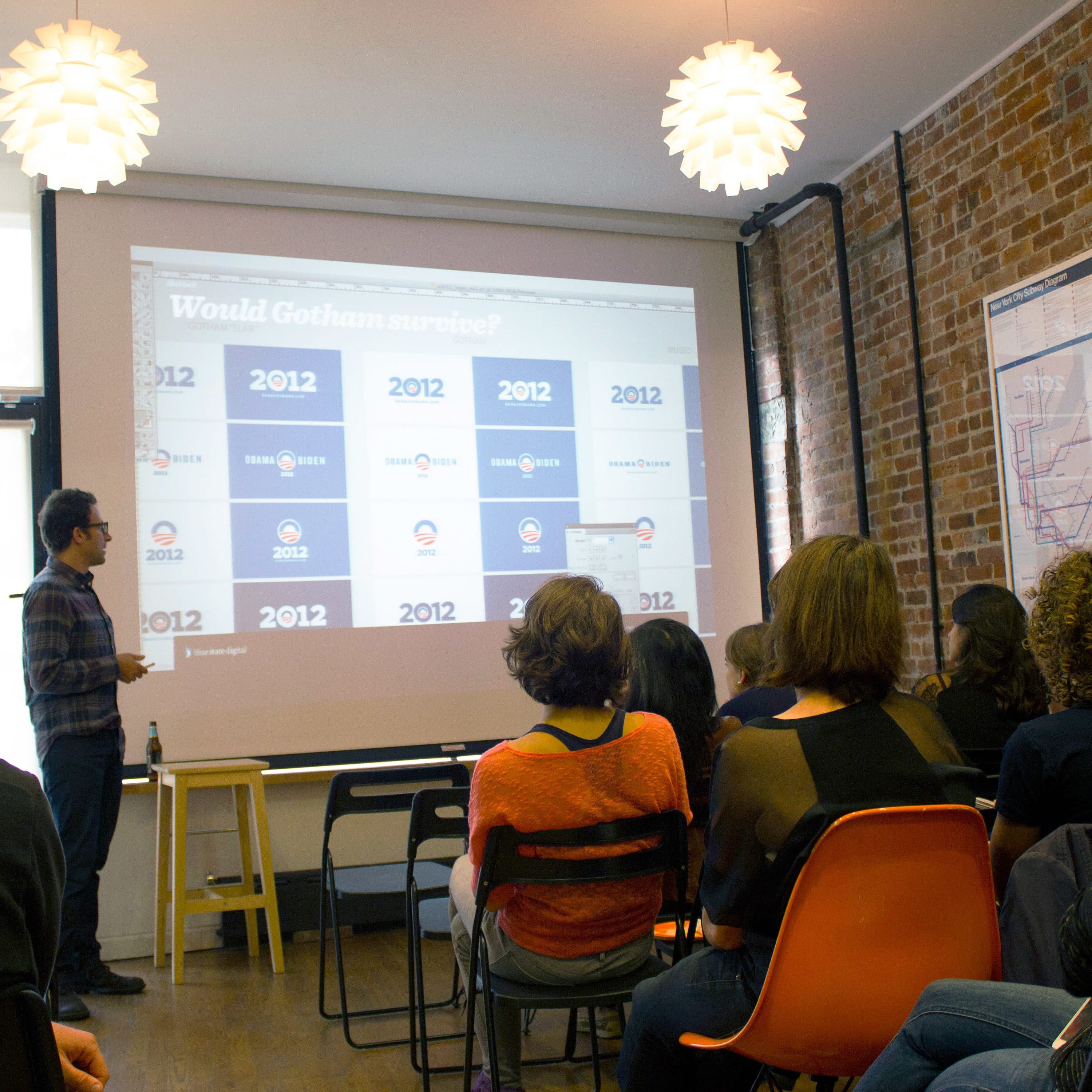 A man presents information displayed on a projected screen to a group of people seated in a room. The room has exposed brick walls, modern pendant lights, and a map on the wall. The audience is attentively watching the presentation.