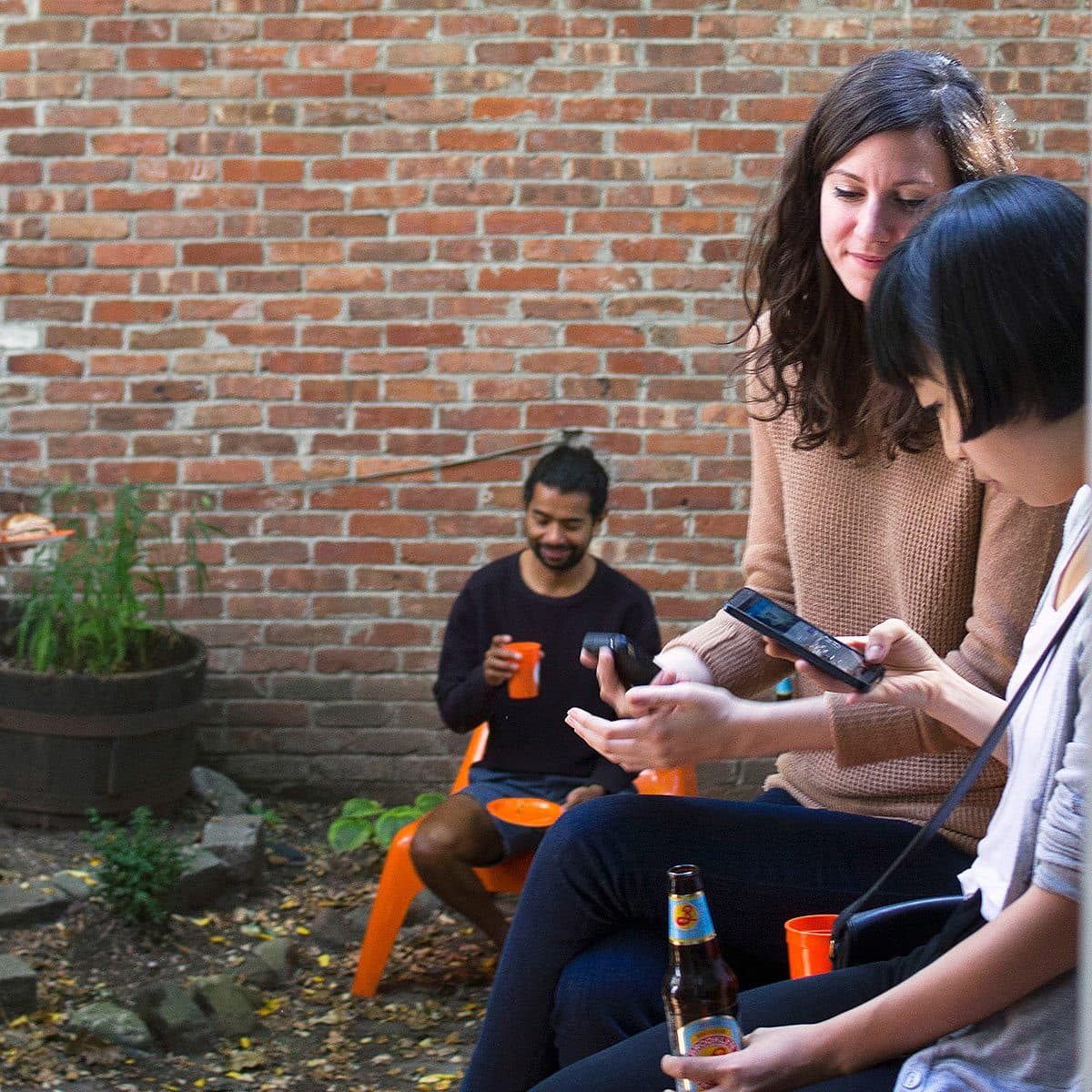 Four people are gathered in a small outdoor area with a brick wall. Two women in the foreground are looking at a phone, and one holds a beer. A man in the background is seated and holding an orange cup, while another man stands nearby with a plate of food.