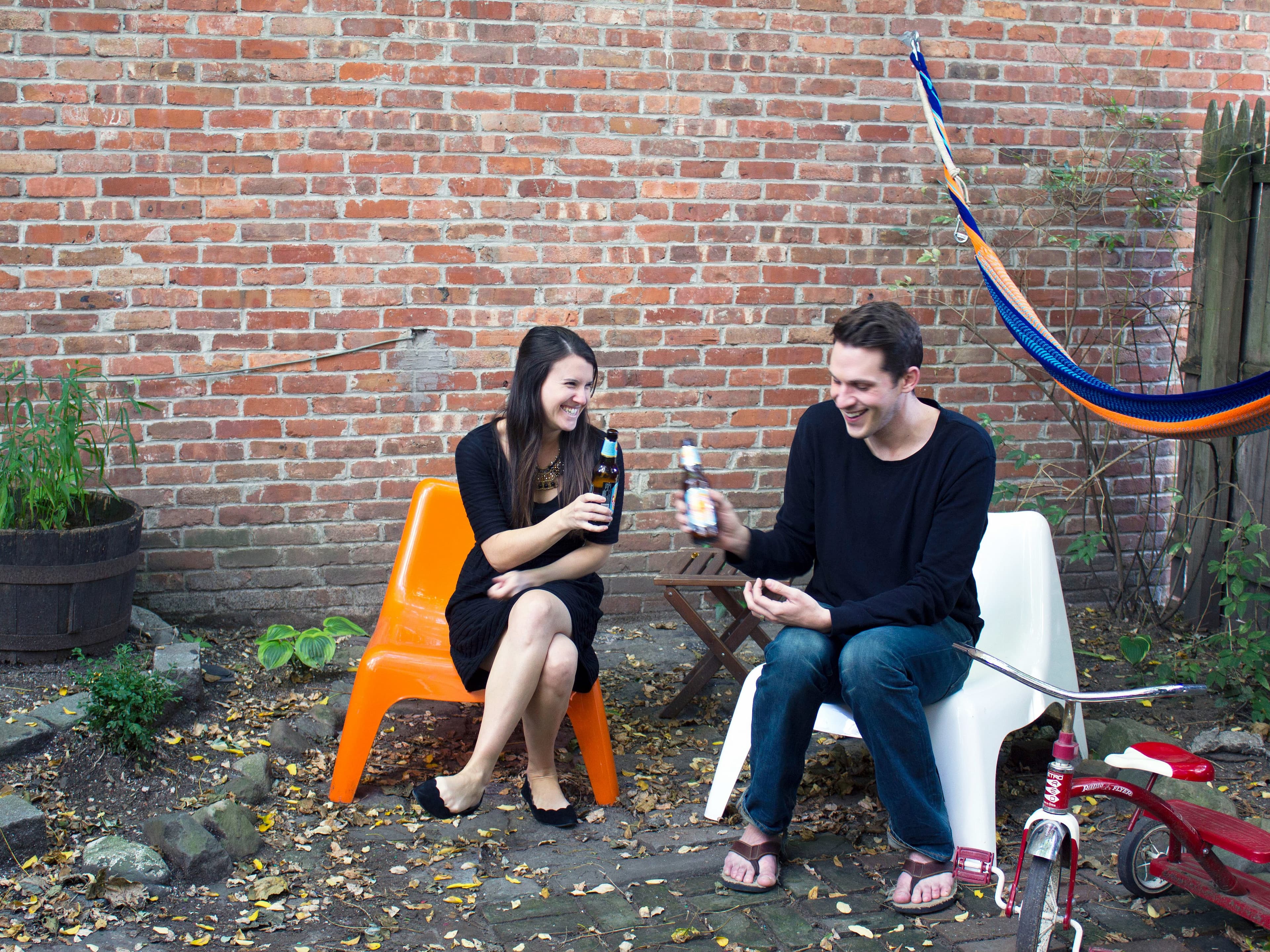 Two people, one in a black dress and one in a black shirt and jeans, are sitting on orange and white chairs in a backyard with a brick wall behind them. They are smiling and holding drinks. There is a hammock, a wooden barrel planter, and a tricycle nearby.