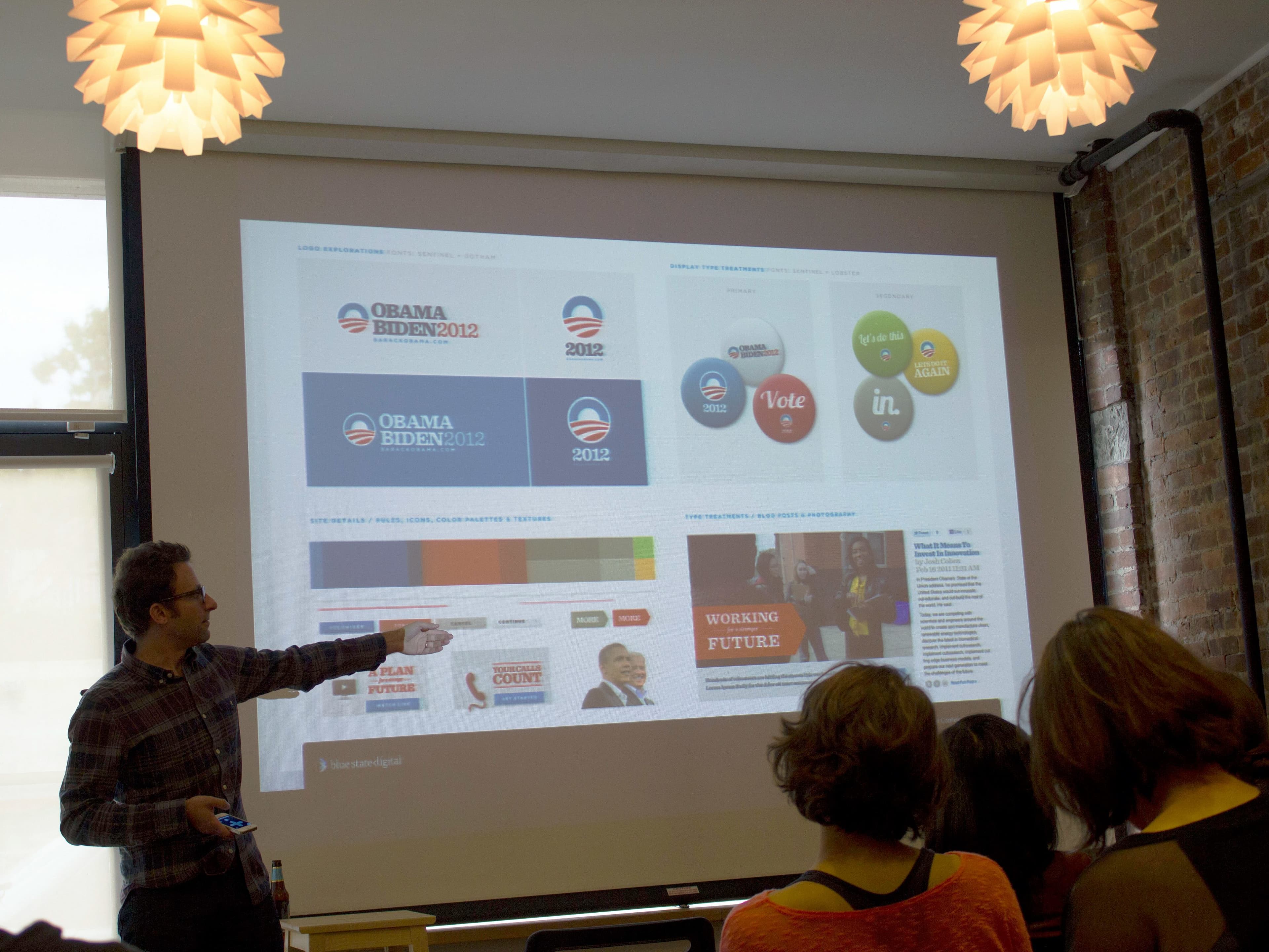A man standing and pointing at a presentation slide projected on a screen in a dimly lit room. The slide displays various Obama-Biden 2012 campaign logos, color palettes, buttons, and print materials. Several people are sitting and watching him attentively.