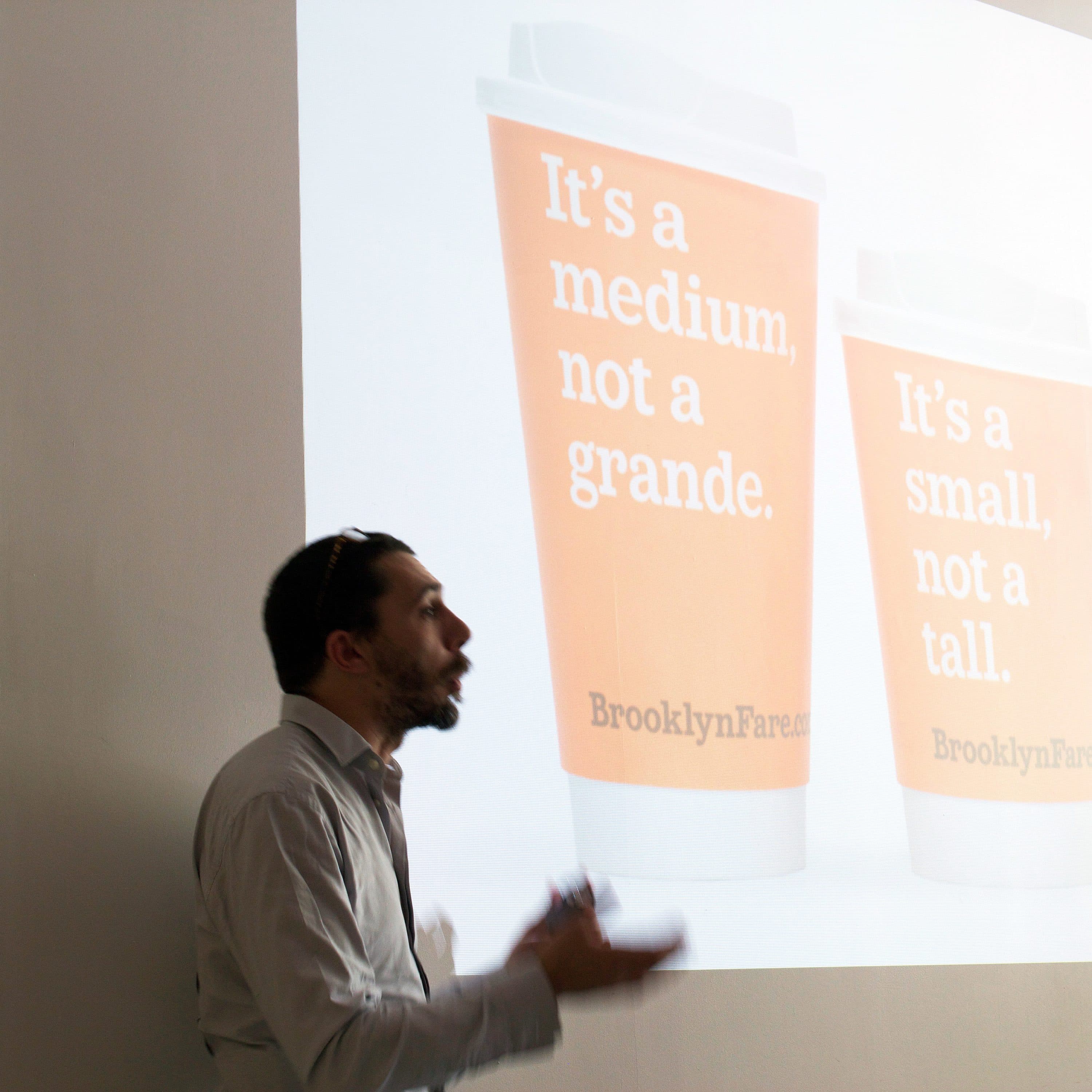 A man gestures in front of a projected image showing two coffee cups. The cup on the left reads "It's a medium, not a grande." and the cup on the right reads "It's a small, not a tall." Both cups display the words "BrooklynFare" at the bottom.
