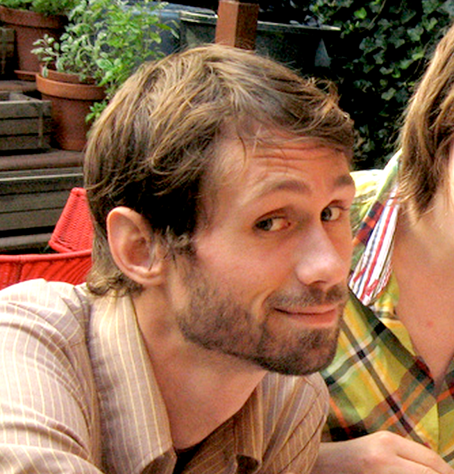 A person with short brown hair and a beard is smiling while looking at the camera. They are outside, with potted plants and greenery in the background. They are wearing a brown shirt, and there is another person next to them, partially visible, wearing a plaid shirt.