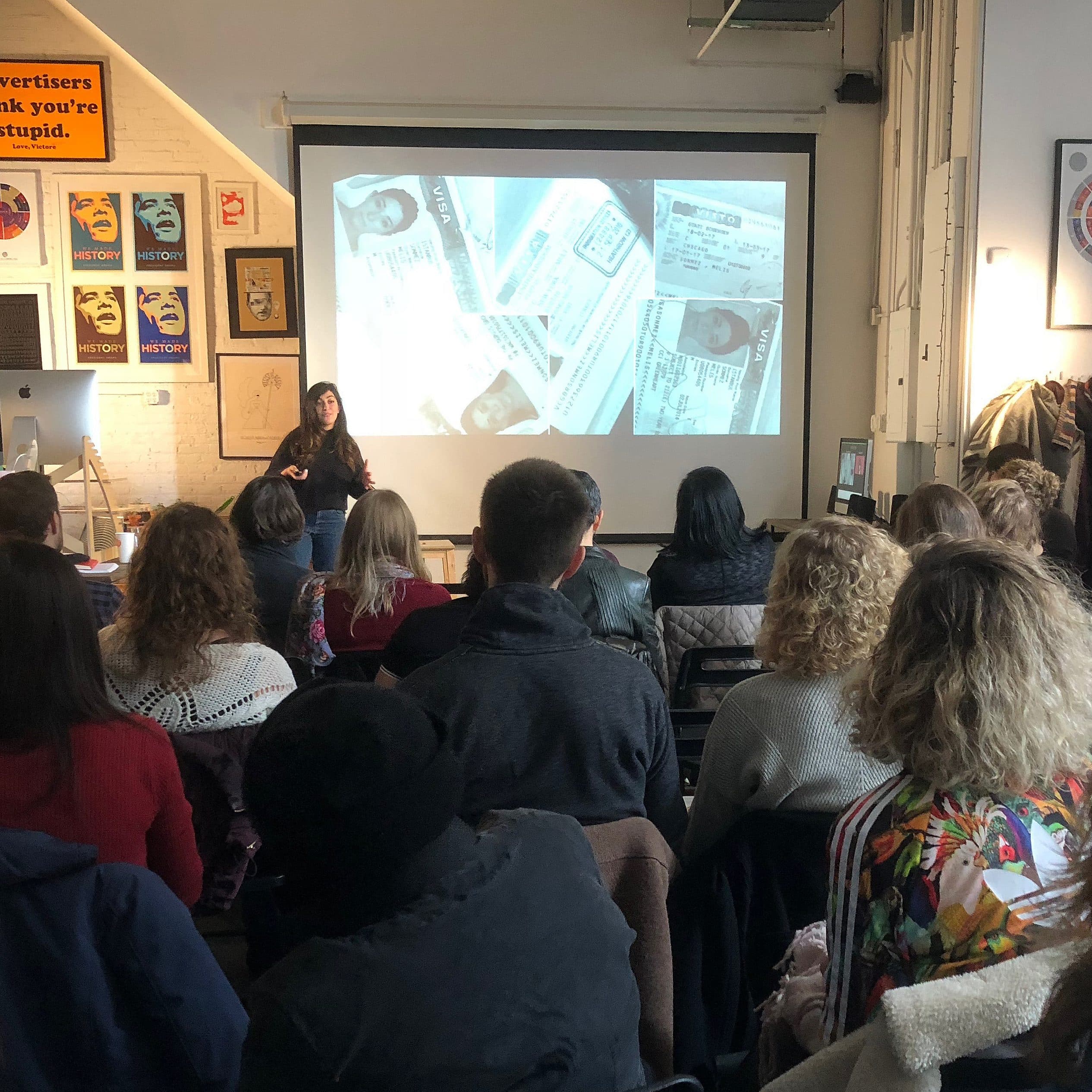 A person delivers a presentation to a seated audience in a modern, stylish room adorned with various posters and artwork. A large projection screen displays a collage of photographs. The audience listens attentively, seated in rows facing the presenter.