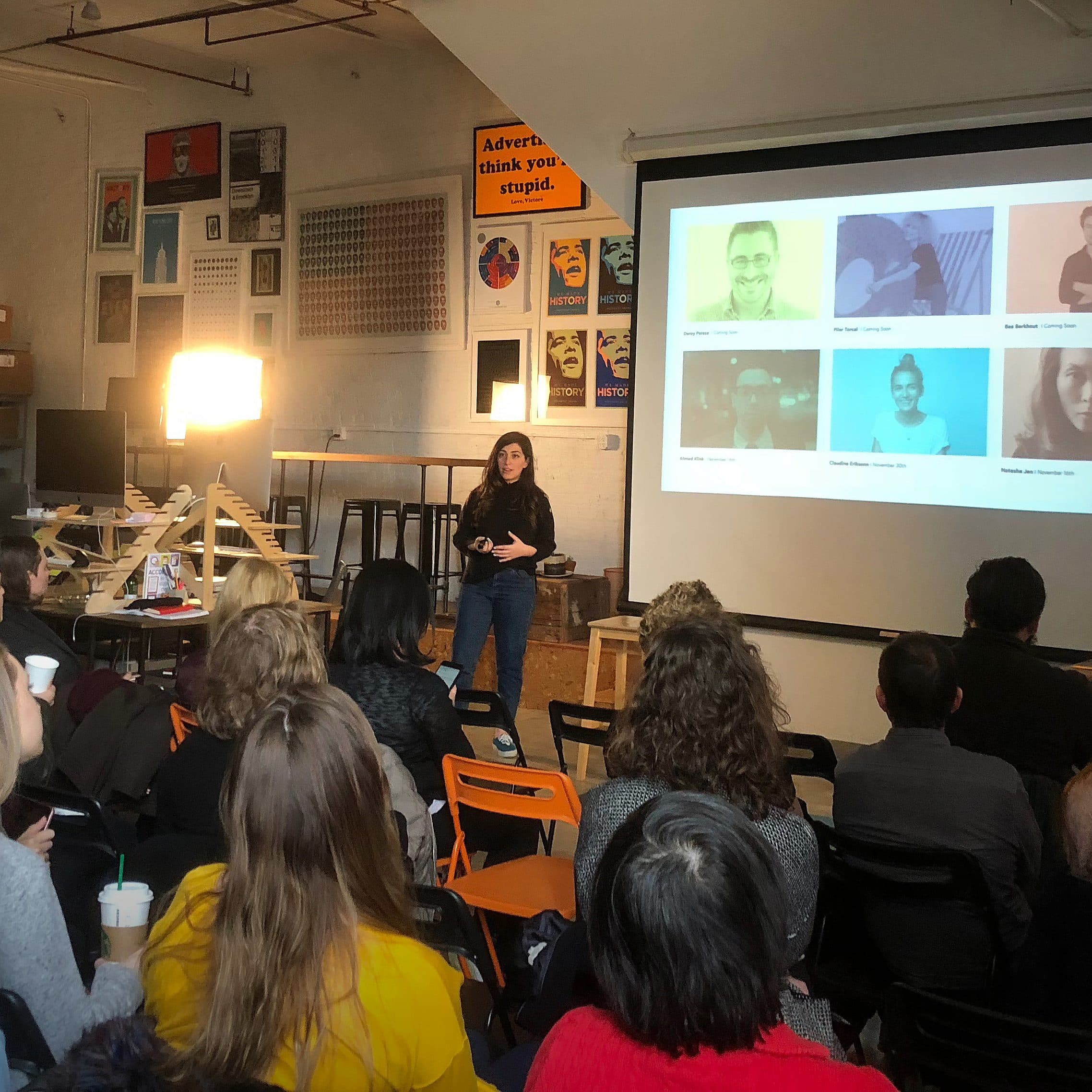 A person stands at the front of a classroom, giving a presentation to a group of seated attendees. The presentation slide shows six individuals with names and titles. The room has a casual setup with various posters and items on shelves.