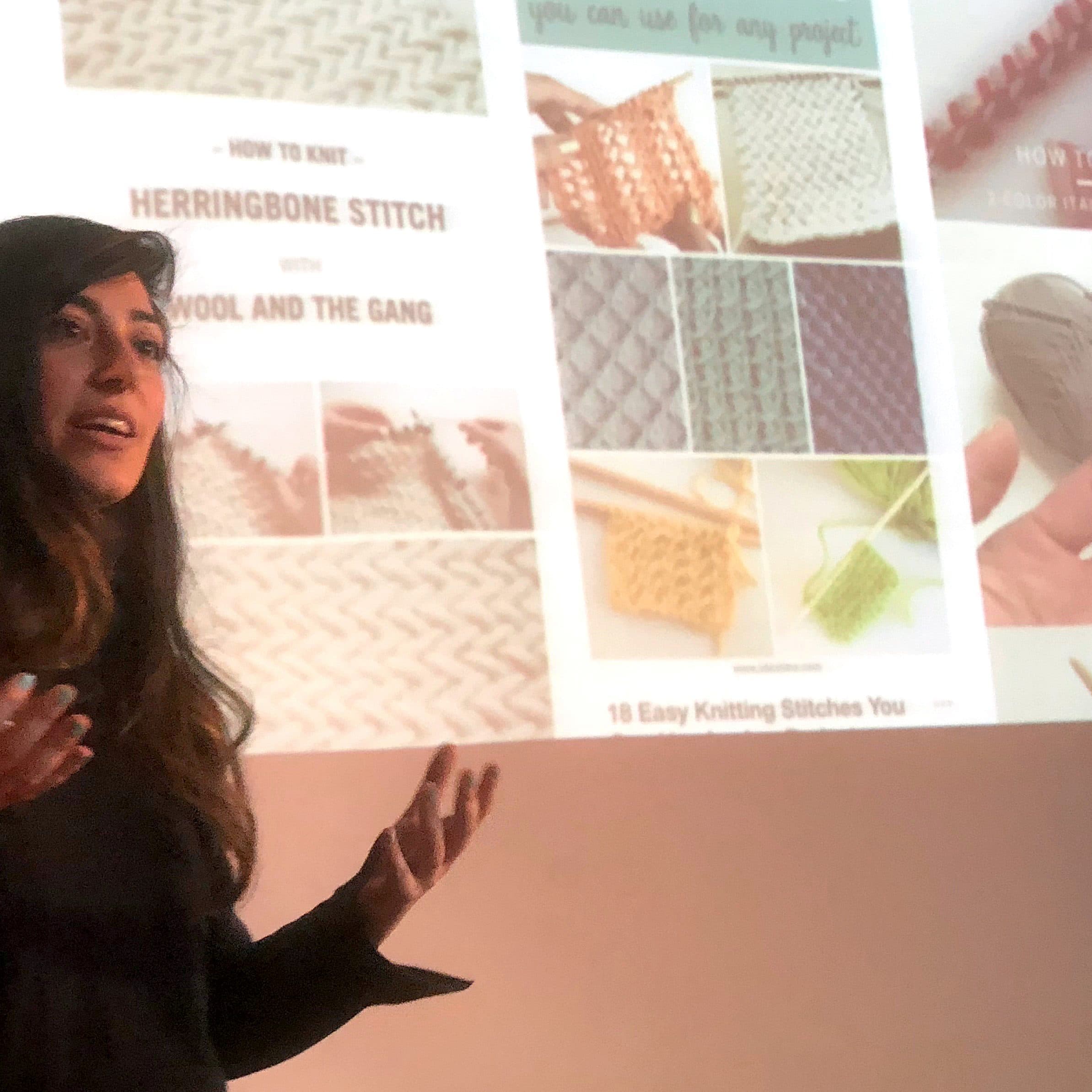 A woman with long dark hair is speaking in front of a screen displaying various knitting stitch techniques and patterns. She is gesturing with her hands, holding a small remote in her right hand.