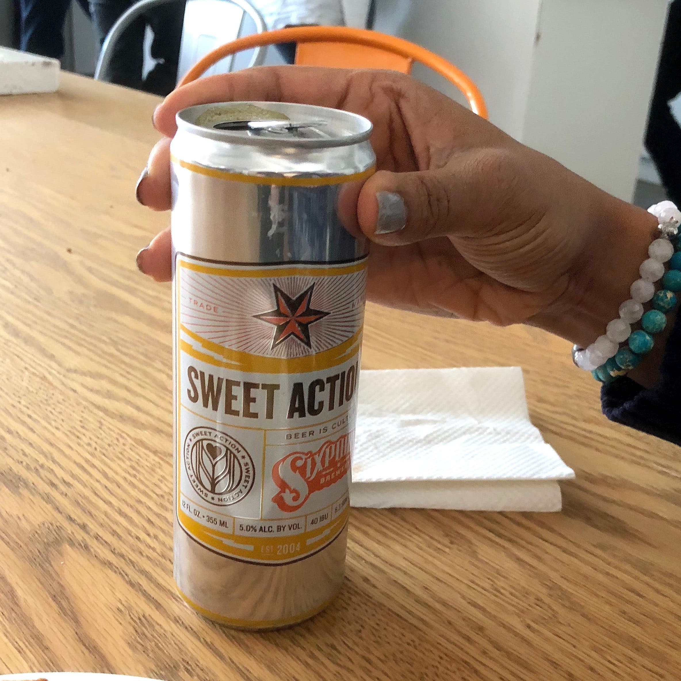 A person holds a can of Sweet Action beer at a wooden table. The can has a label featuring a star and text. A plate with a partially eaten sandwich is in the foreground, and there are people and a kitchen area in the background.