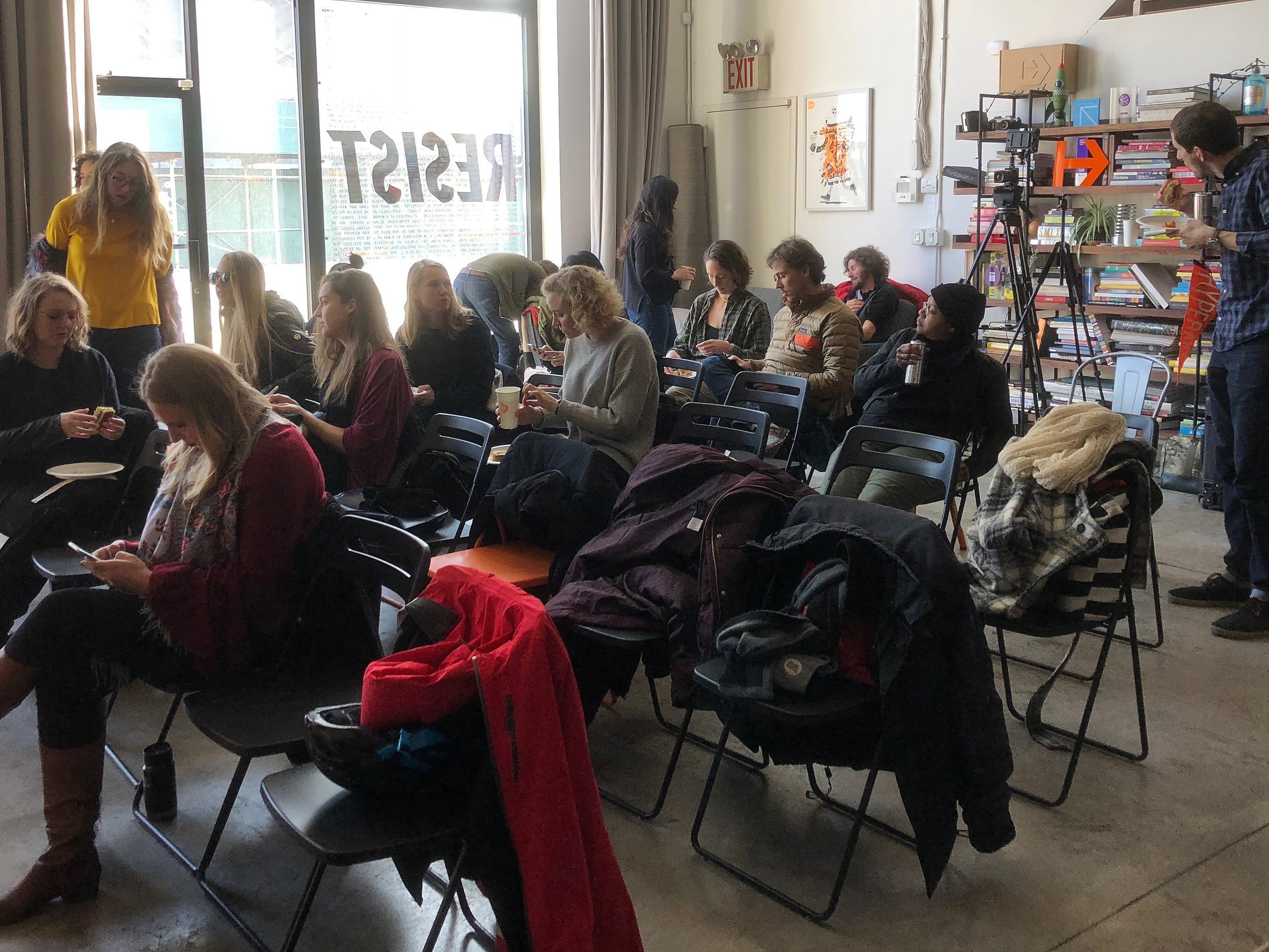 A group of people seated and standing in a room with a bookshelf and a large window. Some people are using cell phones, while others are talking. Jackets and coats are draped over chairs. The word "RESIST" is visible on the window in large letters.