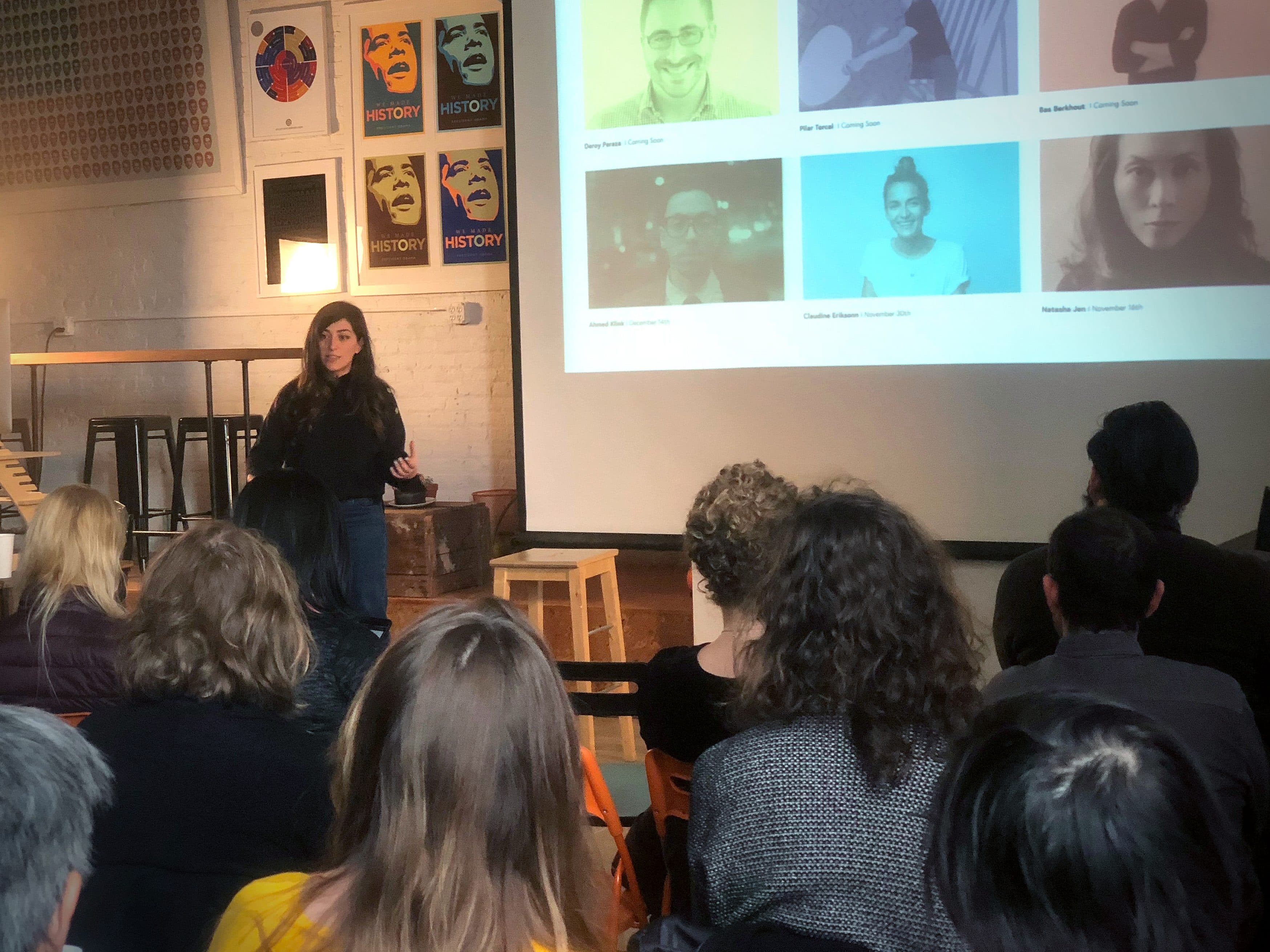 A woman is speaking in front of an audience in a classroom or workshop setting. Behind her is a large screen displaying various headshots and names. The audience is seated facing her, and there are posters on the wall with the word "HISTORY.