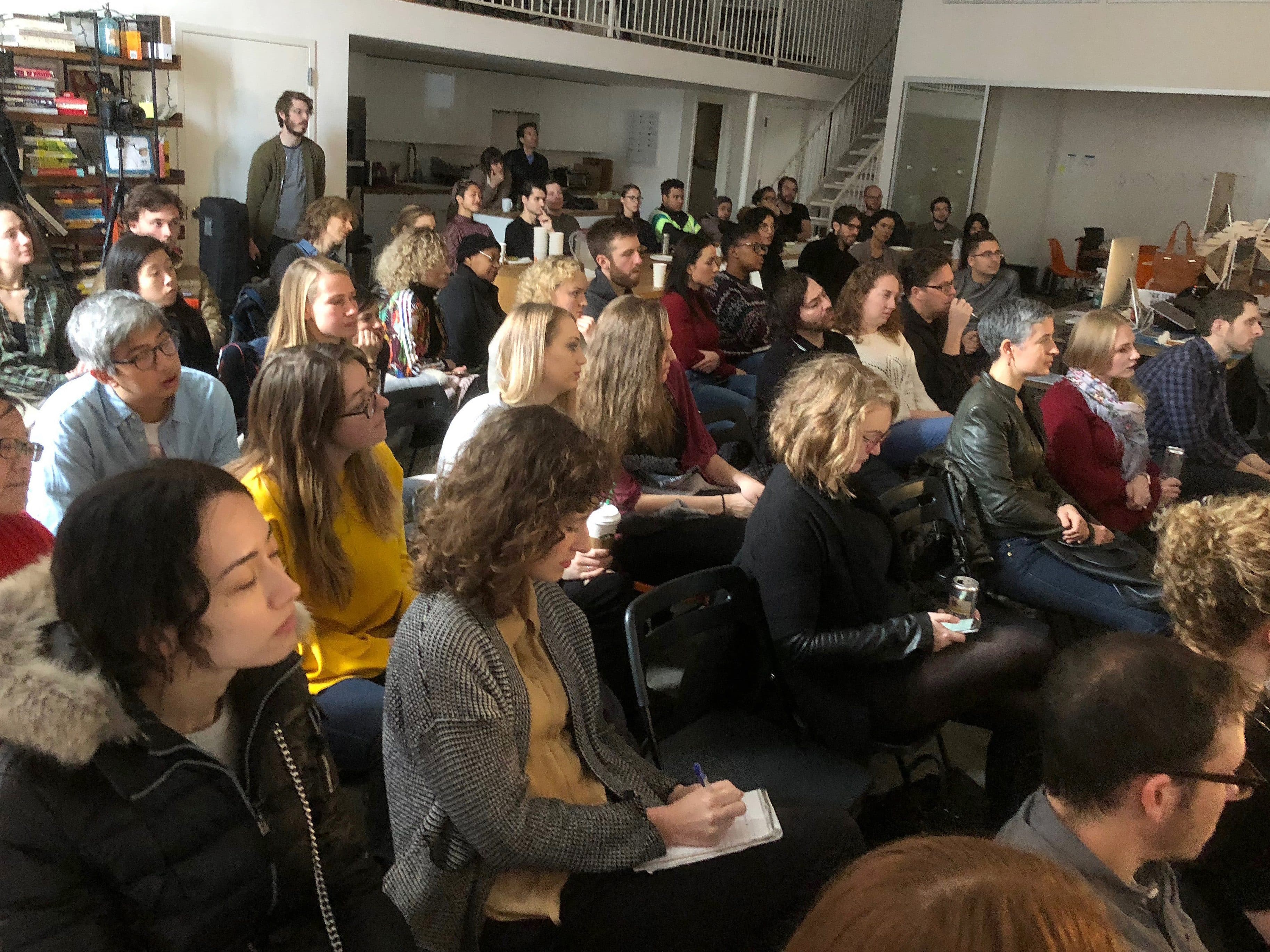 A diverse group of people sitting closely together in a well-lit room, attentively listening to a presentation. Some individuals are taking notes, while others are holding drinks. The room has bookshelves and a staircase in the background.