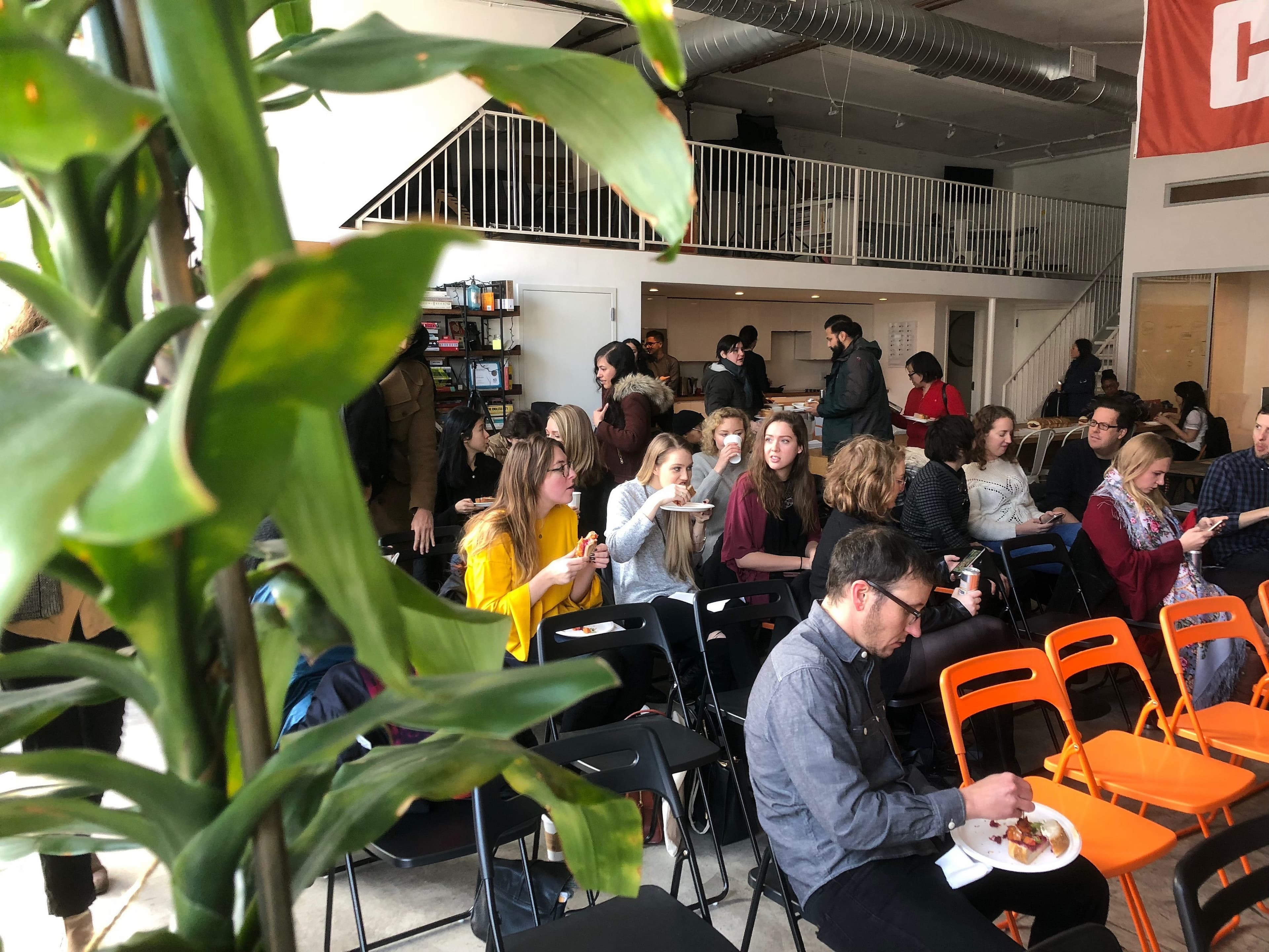 A group of people sit on black and orange chairs in a modern, spacious room with a loft area. They are socializing and eating, creating a lively atmosphere. In the foreground, a large green potted plant adds a touch of nature to the scene.