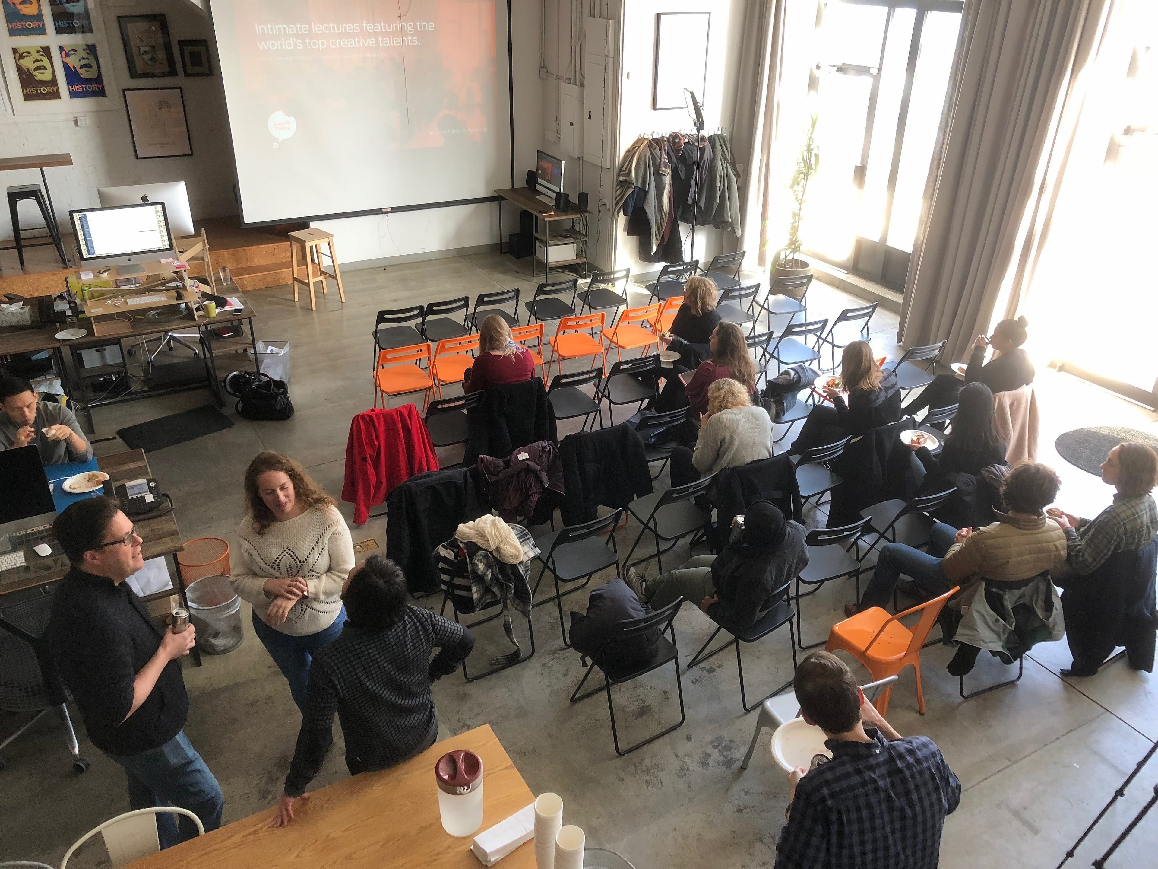 A group of people are gathered in a bright room with a projector screen at the front displaying a presentation. There are chairs set up in rows, and some people are standing and chatting while others are seated. The room has a modern, casual atmosphere.