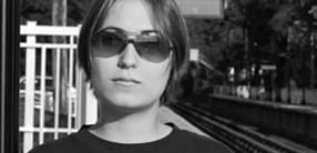 A person with short hair and wearing sunglasses stands in front of a train platform. The image is in black and white. The individual looks directly at the camera with a neutral expression. The background includes a railway track and some trees.