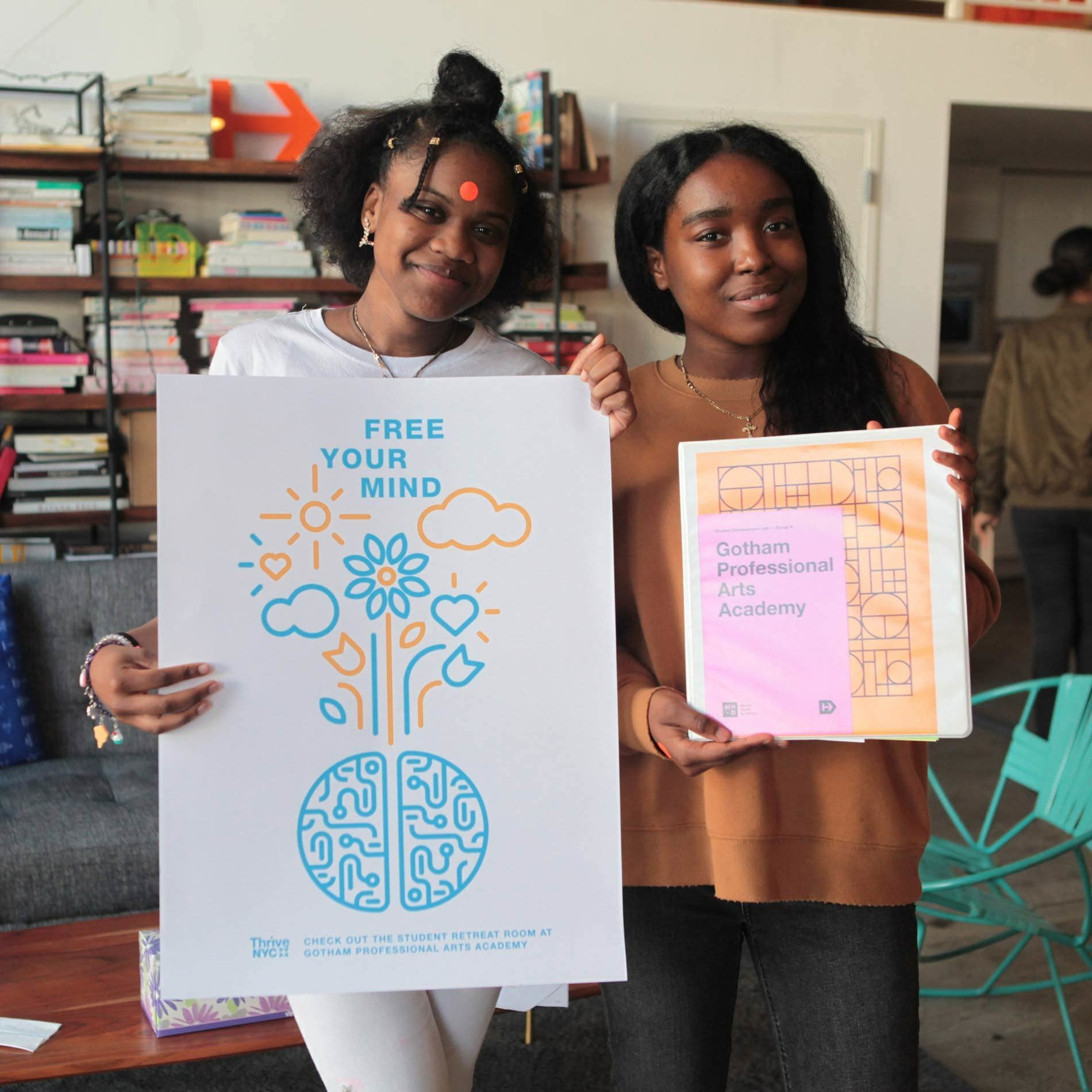 Two smiling young women stand side by side, holding posters. The poster on the left reads "Free Your Mind" with a drawing of a brain and flowers. The poster on the right reads "Gotham Professional Arts Academy" with a geometric pattern. A bookshelf is in the background.