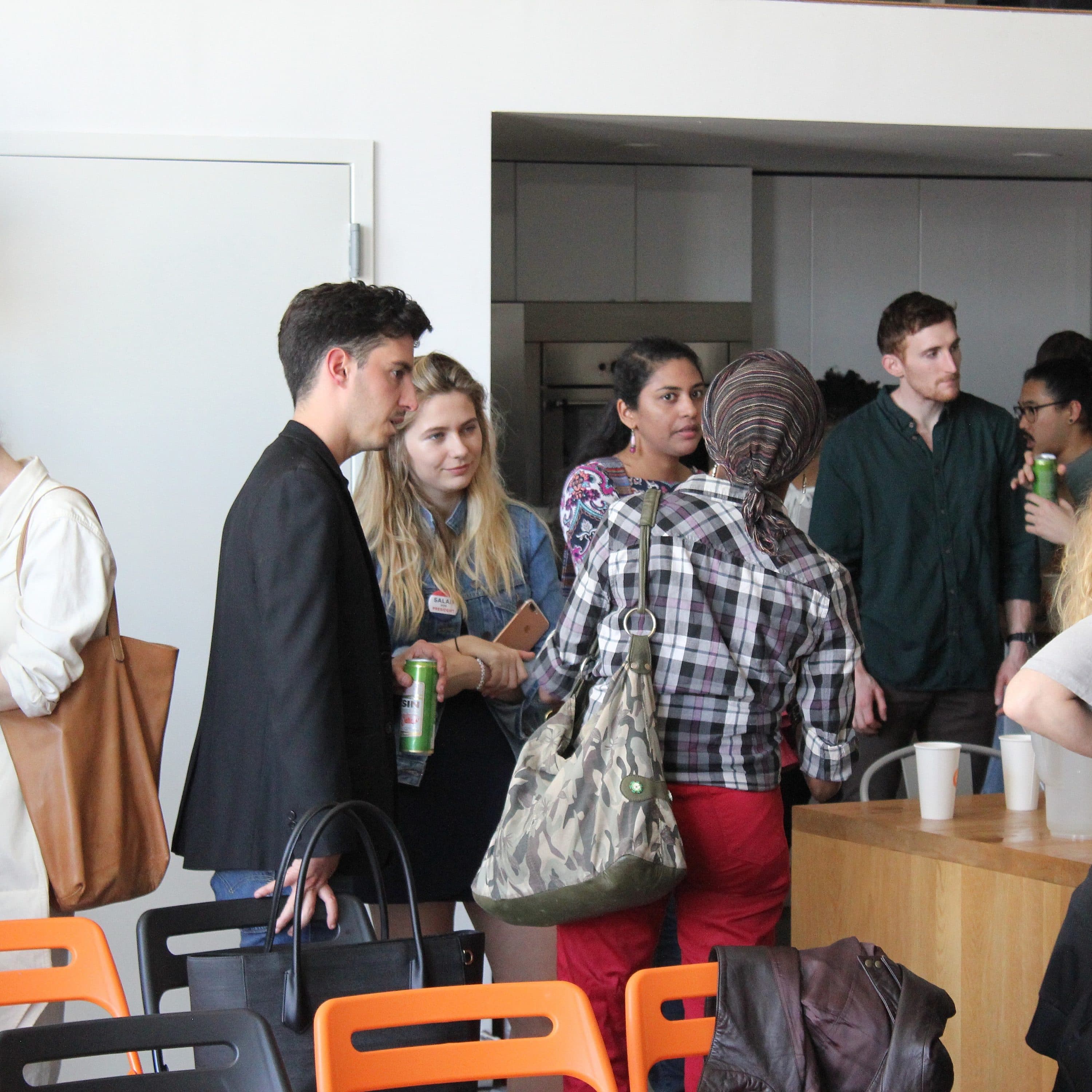 A group of people is gathered in a casual setting, chatting and socializing. Some hold drinks while standing near a counter. The background features shelves with books and various items. There are orange and black chairs visible in the foreground.