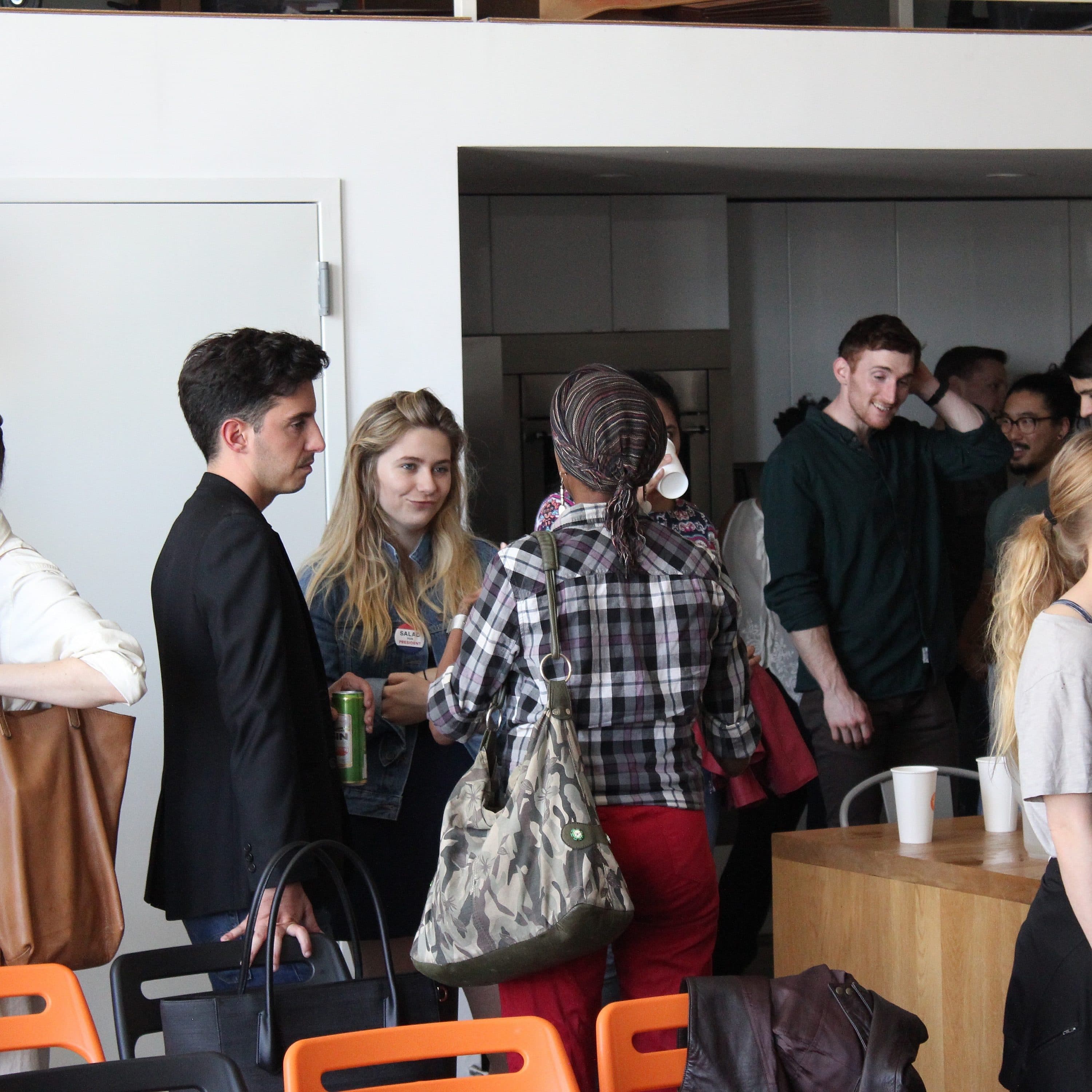 A group of people are gathered in a modern indoor space with shelves and a door visible in the background. Some people are engaged in conversation, while others seem to be waiting or looking around. There are bright orange chairs and a wooden table visible.