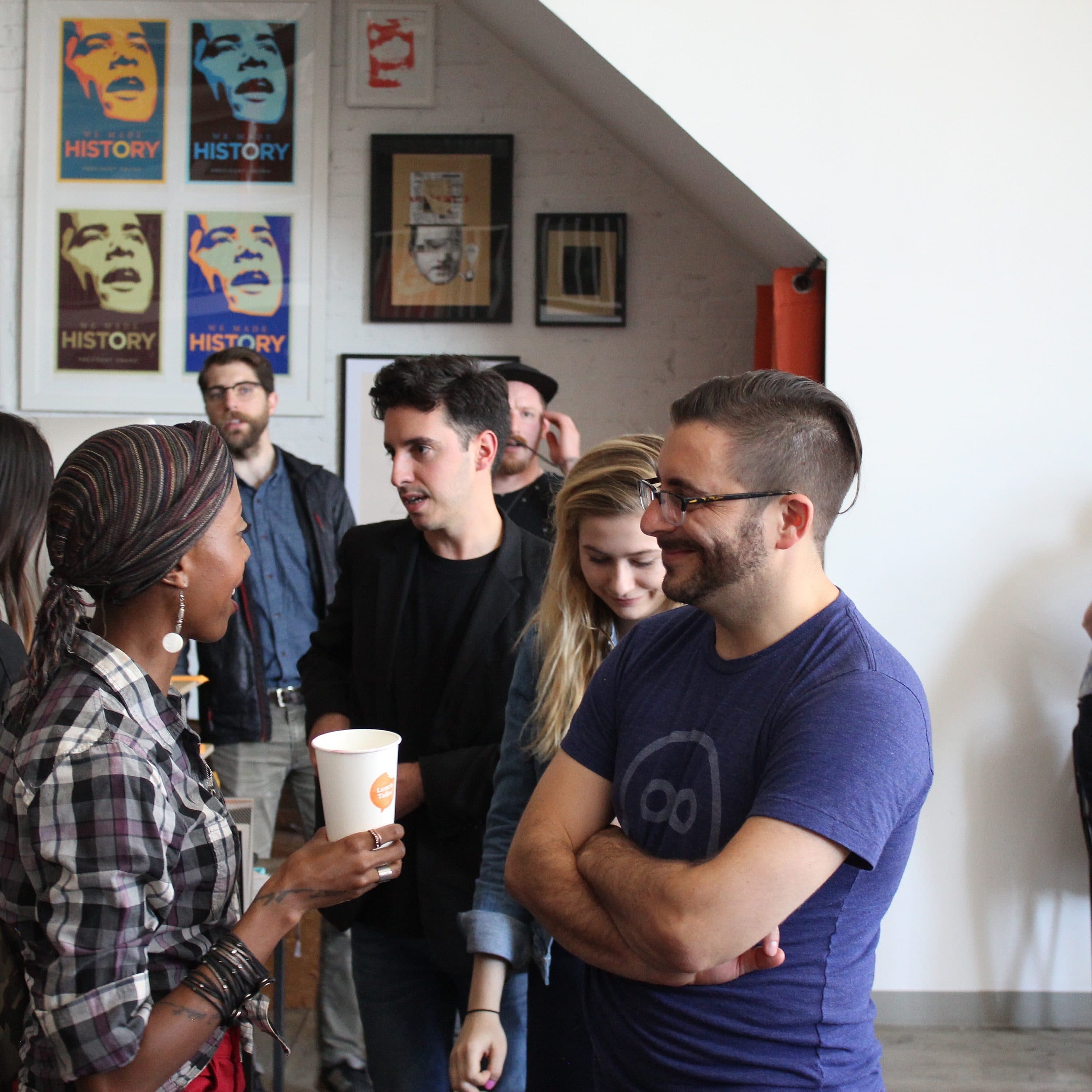 A group of people are conversing in a casual setting. One person holds a coffee cup while talking to another who has arms crossed. The background displays various posters, including some with the word "HISTORY," and a subway map. A person is bending over a table.