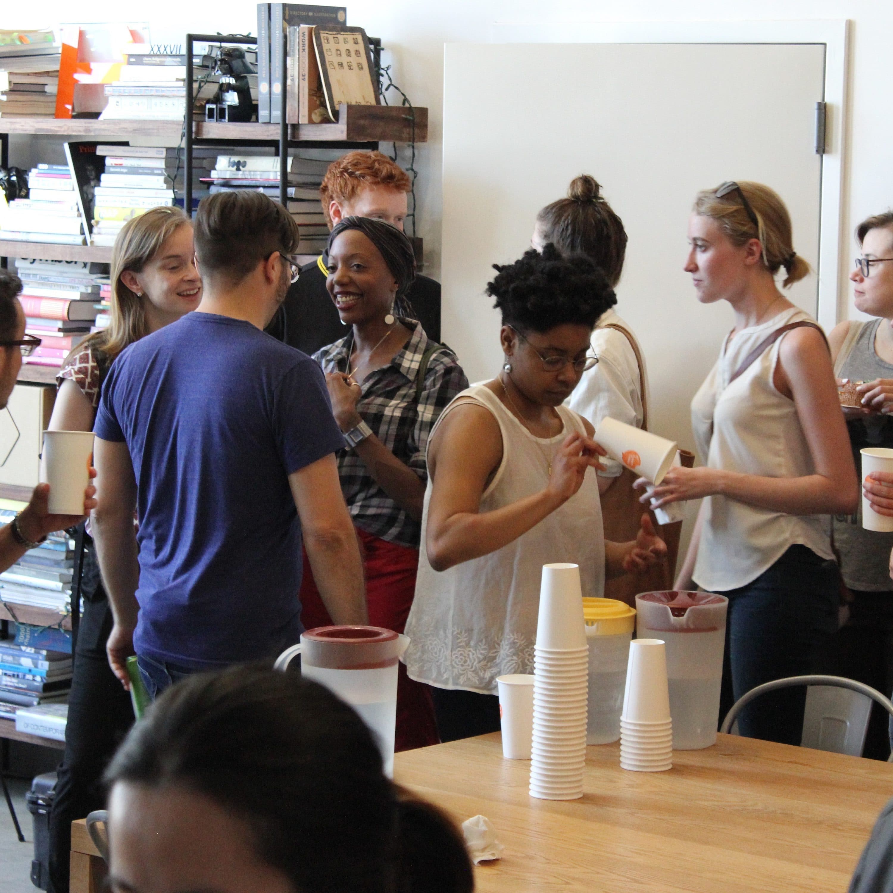 A group of people are gathered in a room, engaged in conversation and serving themselves from pitchers on a table with stacked cups. Bookshelves filled with books and a camera are visible in the background. The atmosphere appears casual and social.