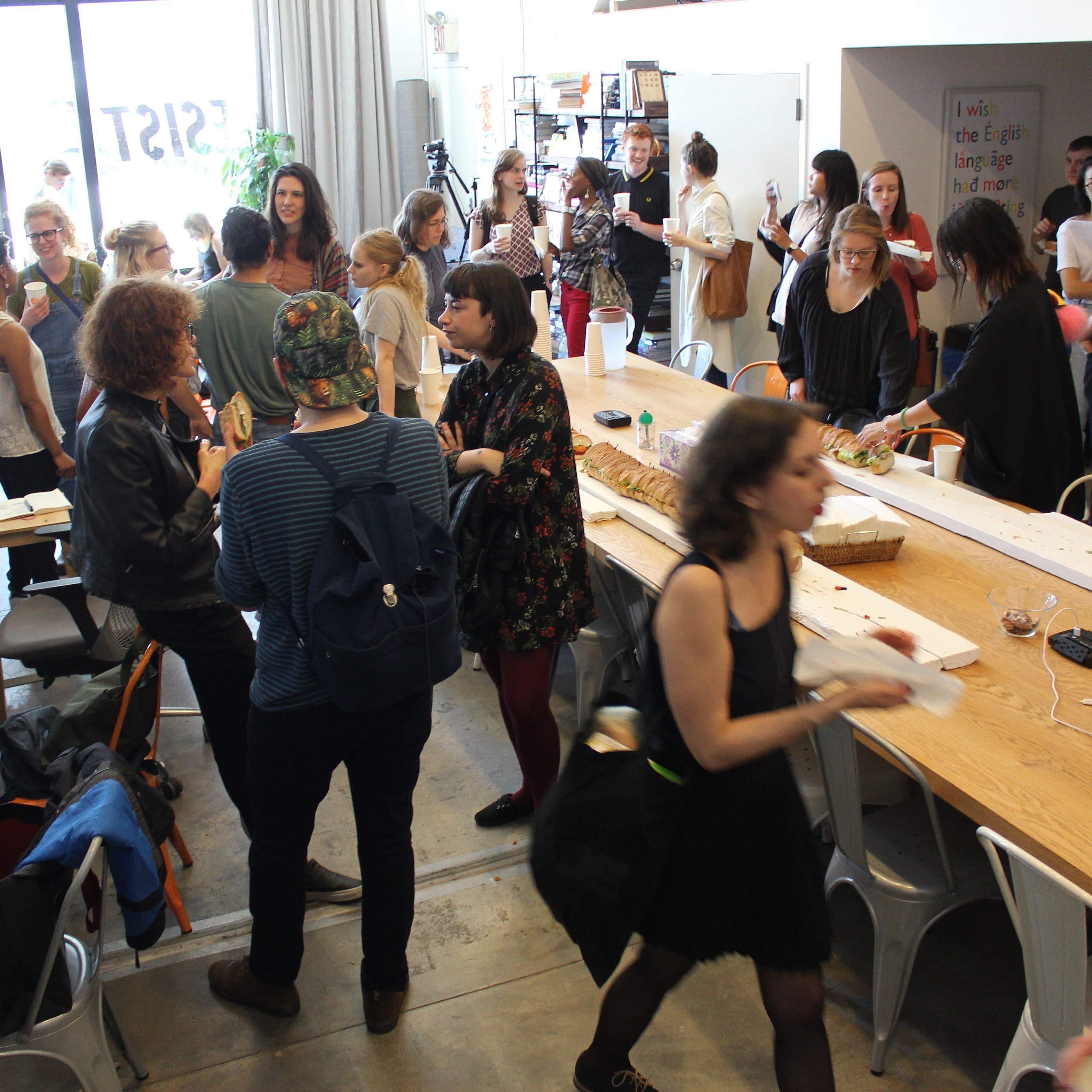 A busy communal workspace with numerous people engaged in conversation and activities. The room features long wooden tables with chairs, assorted items, and office supplies. Natural light streams in from windows, and various shelves and workstations can be seen in the background.