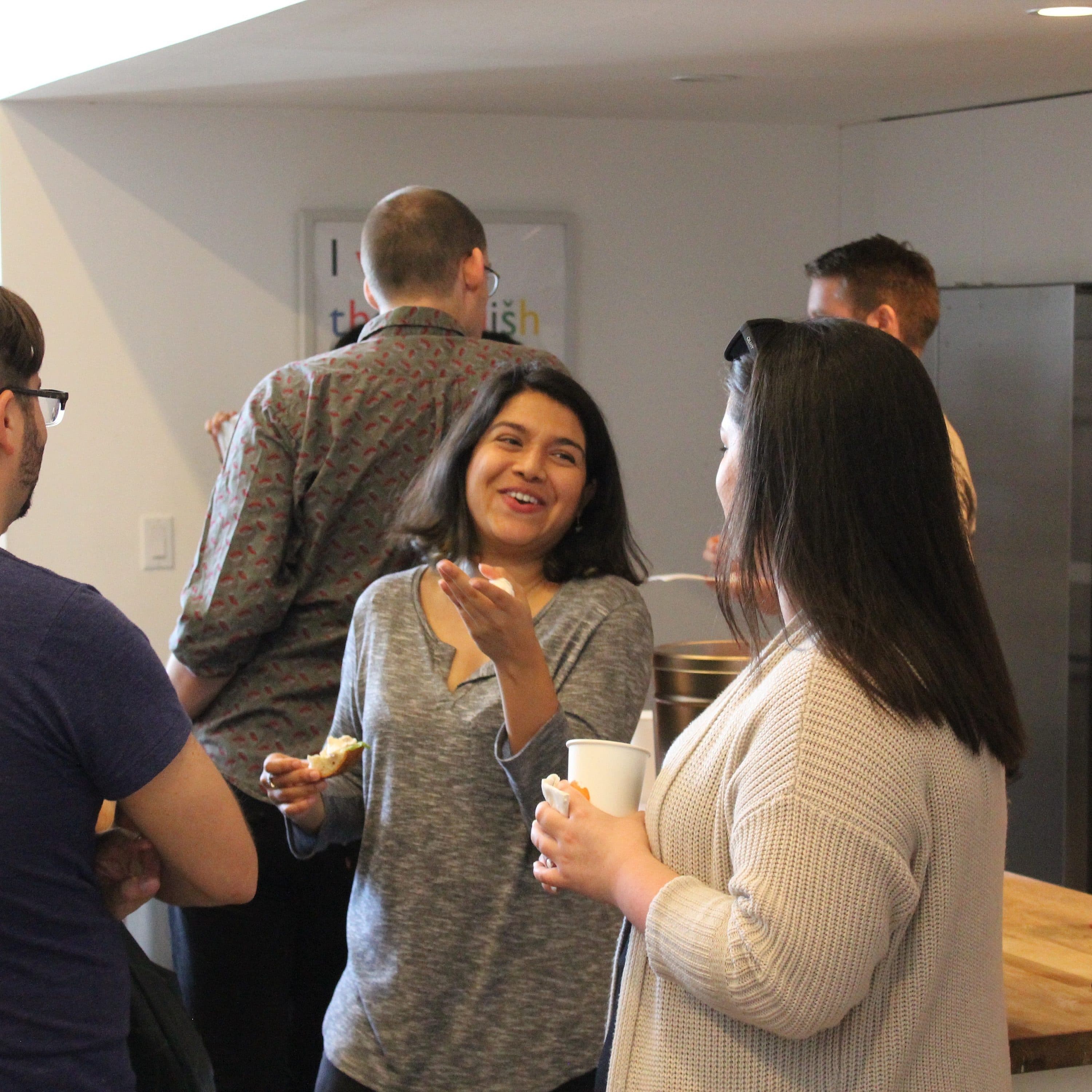 A group of people are gathered in a kitchen area, engaging in conversation. Some hold cups, and one woman in the center is smiling and gesturing with her hand. The environment appears casual and friendly, with a countertop and kitchen appliances in the background.