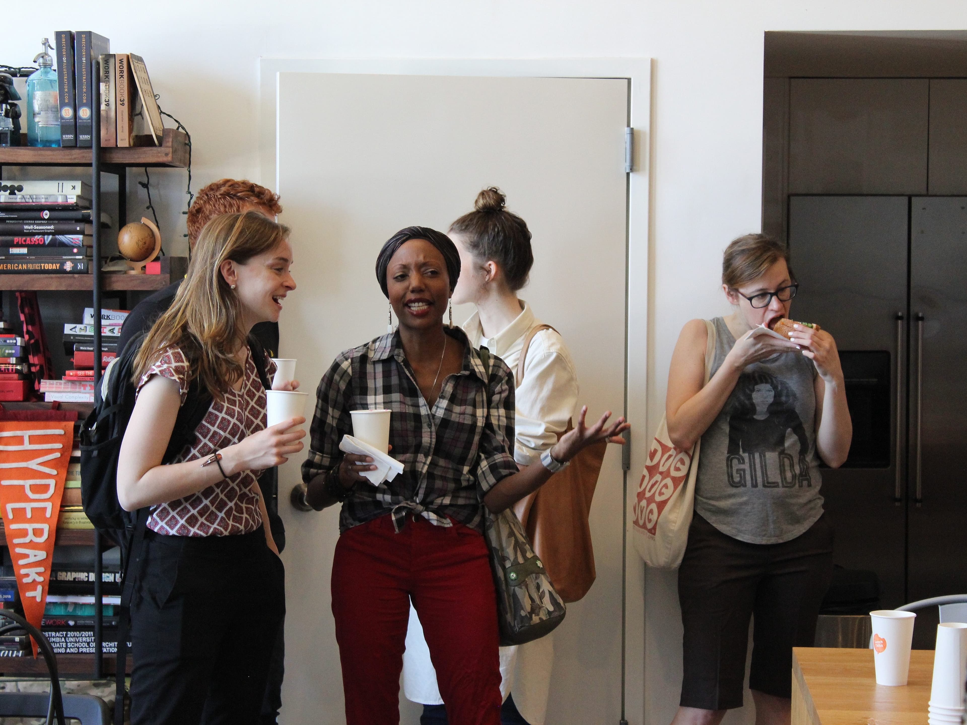 A group of people converse in a casual, indoor setting with a bookshelf in the background. Four people are visible; one is eating, two are holding coffee cups, and another gestures while talking. The atmosphere appears relaxed and friendly.