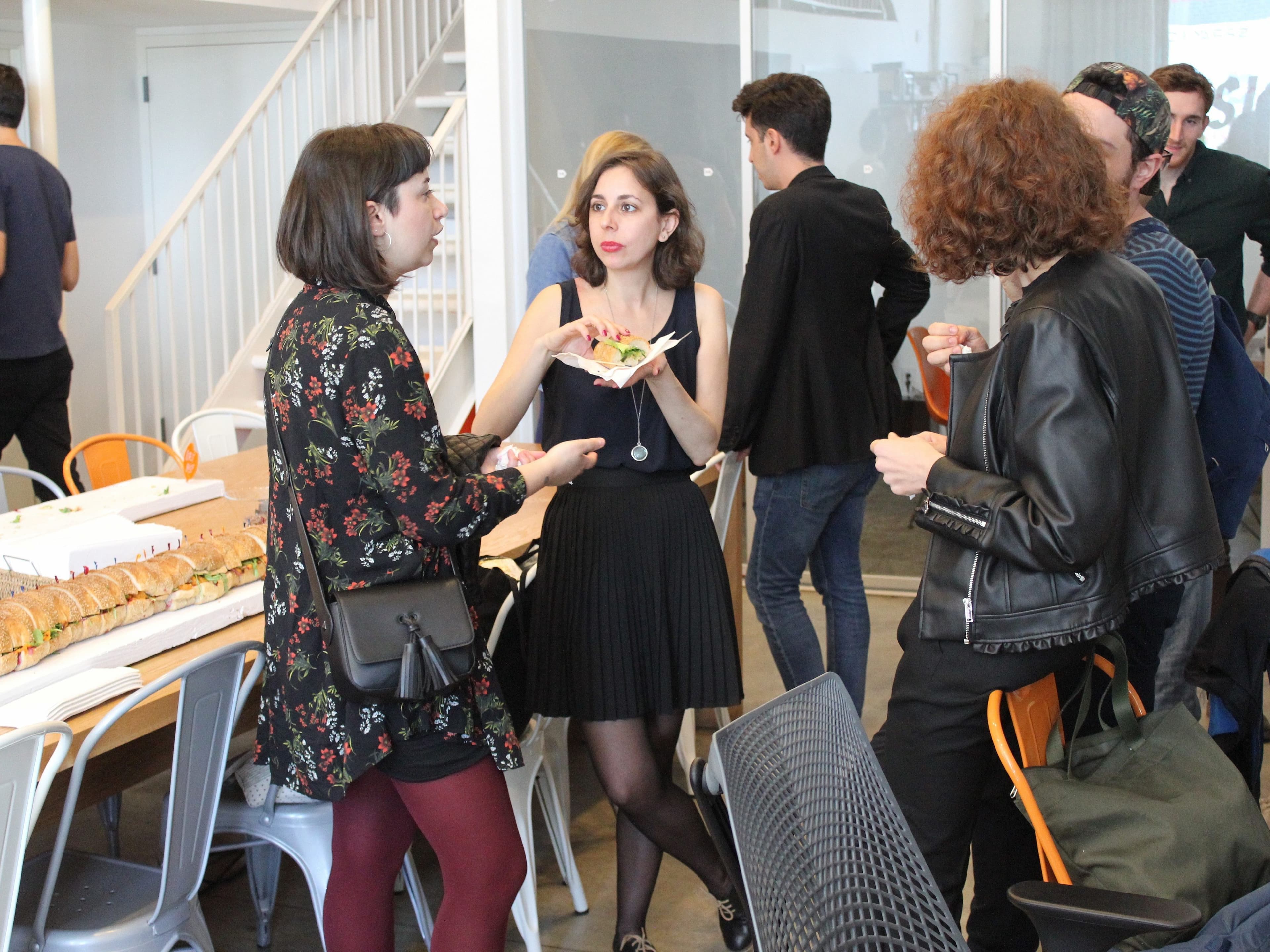 A group of people socialize in a casual indoor setting. Three women stand in the foreground, one holding a plate of food. Others are mingling or standing near a long table with a large sandwich. The space has modern decor with white walls and chairs.