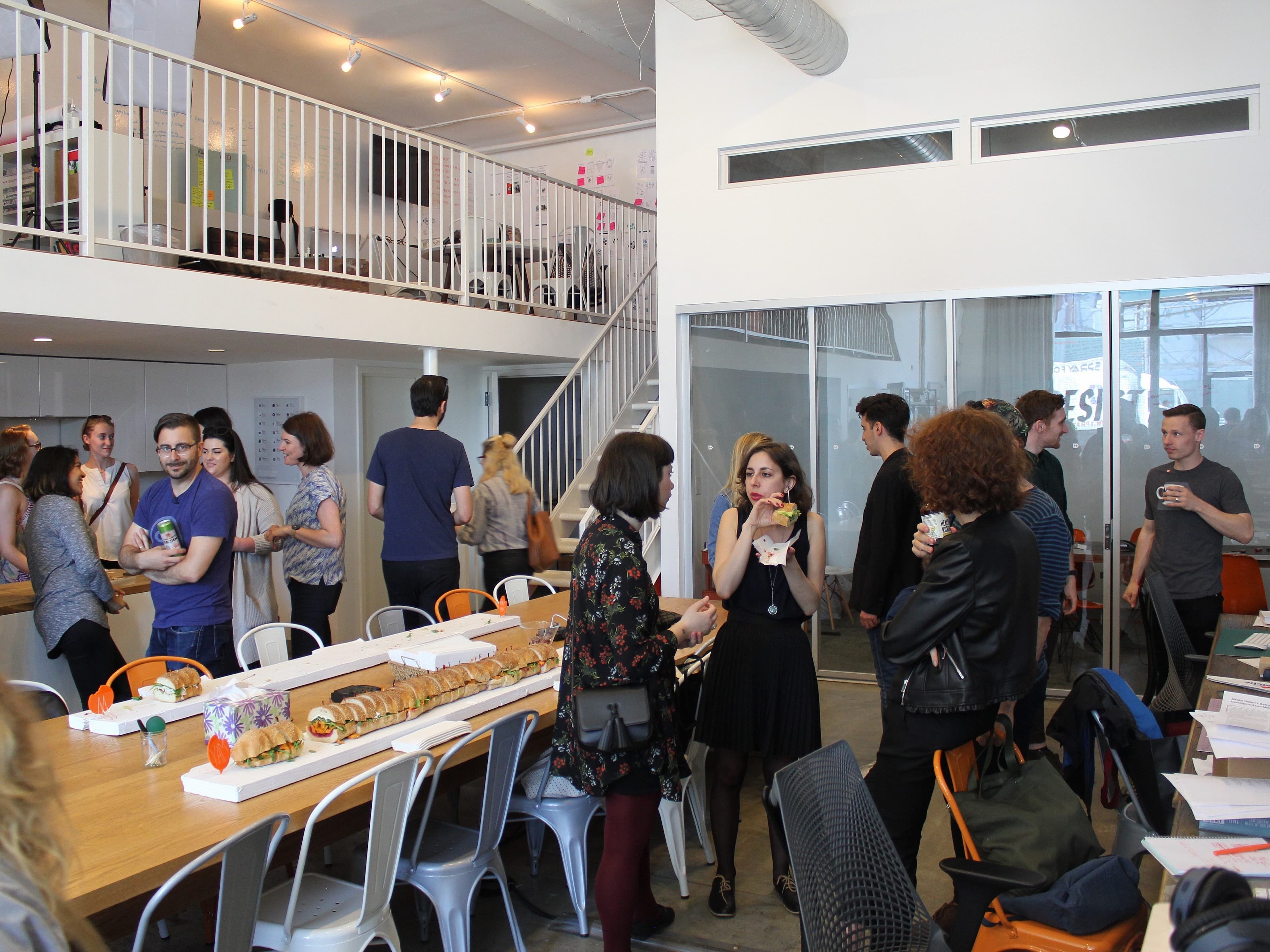 A group of people are gathered in a modern, open-space office. Some are chatting, while others are enjoying refreshments. There's a long table with sandwiches in the foreground, tables with papers, and a staircase leading to a mezzanine level in the background.